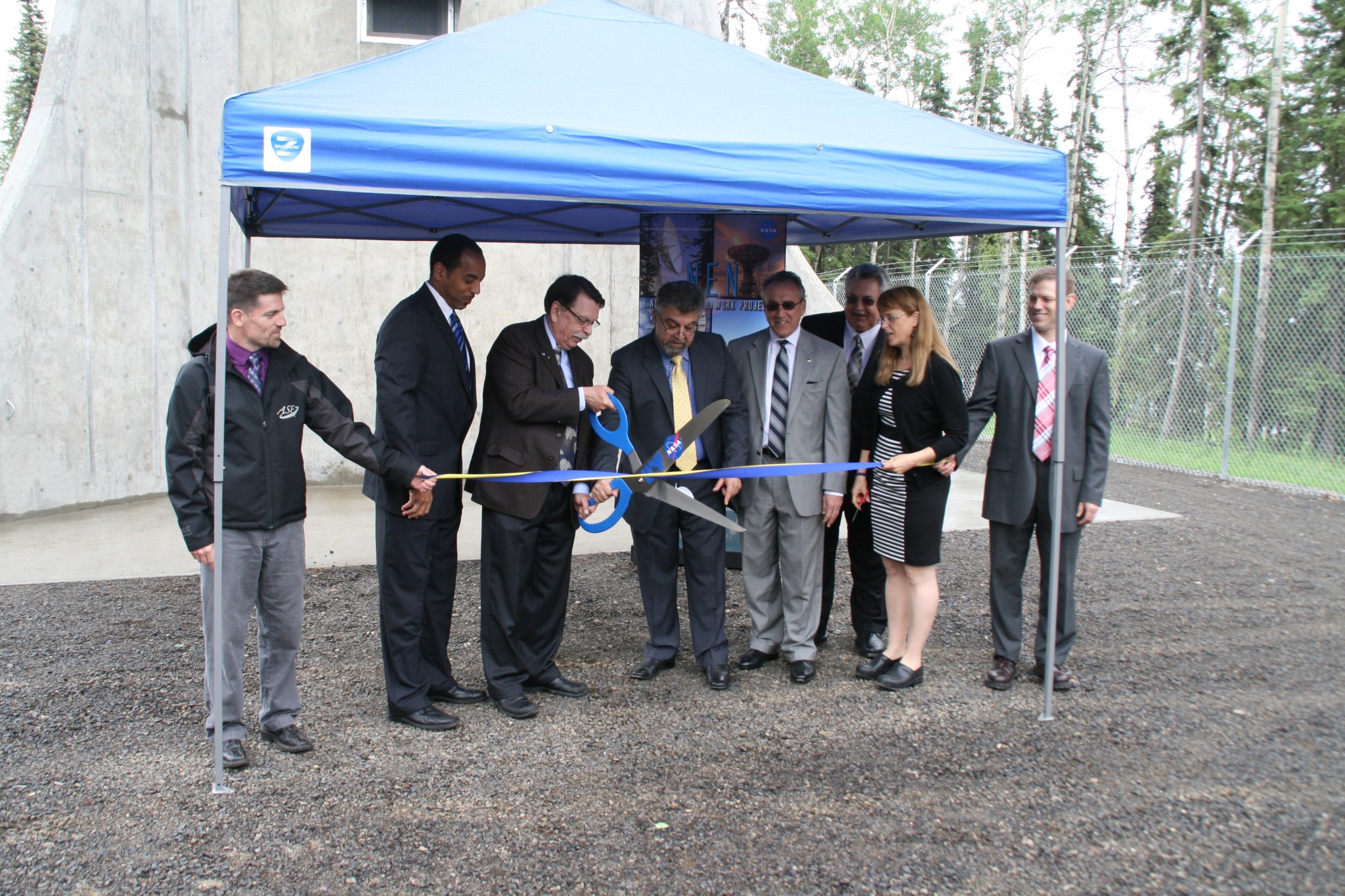 7 men and one woman at a ribbon cutting ceremony in the rain under a blue tent. 