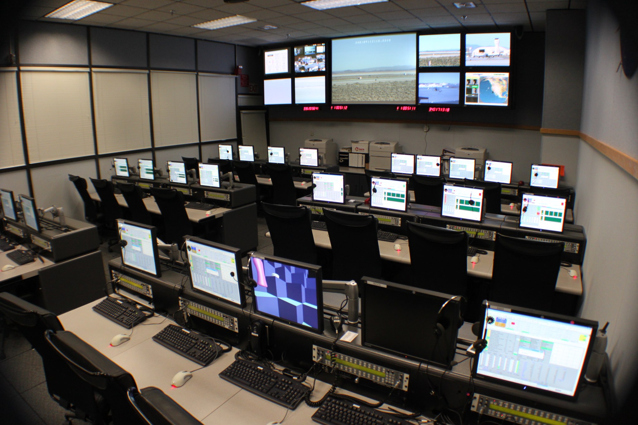 A room of tables with multiple stations, each with monitors and keyboards and a large screen at the front of the room with multiple monitors.