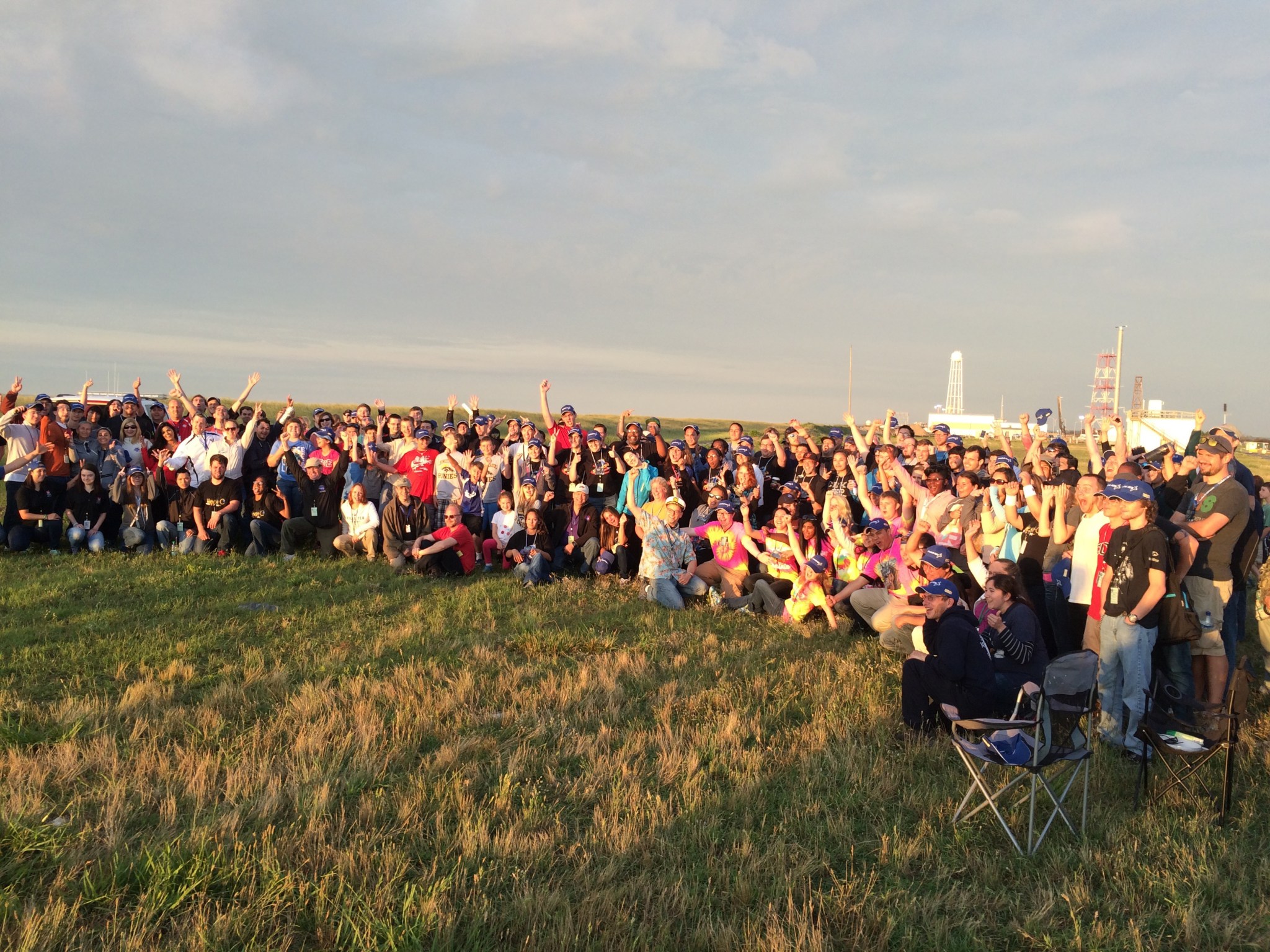 A large group of more than 100 students poses for a group photo in a field in front of a rocket launch.