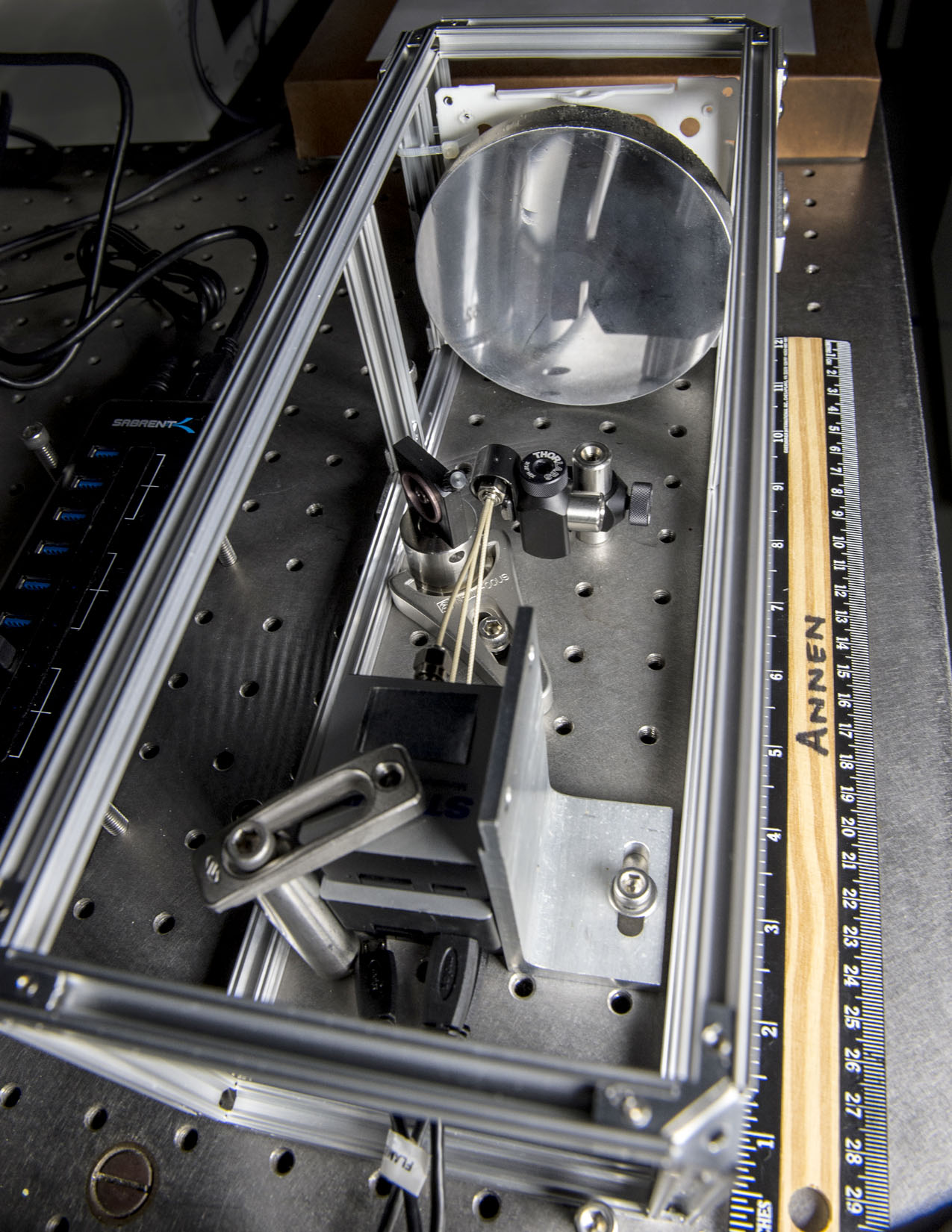 A mechanical instrument made of a silver metal frame. In the frame, a silver mirror is propped up on one end. Other components are drilled into a board under the frame. A ruler next to the frame shows that it's about one foot long.