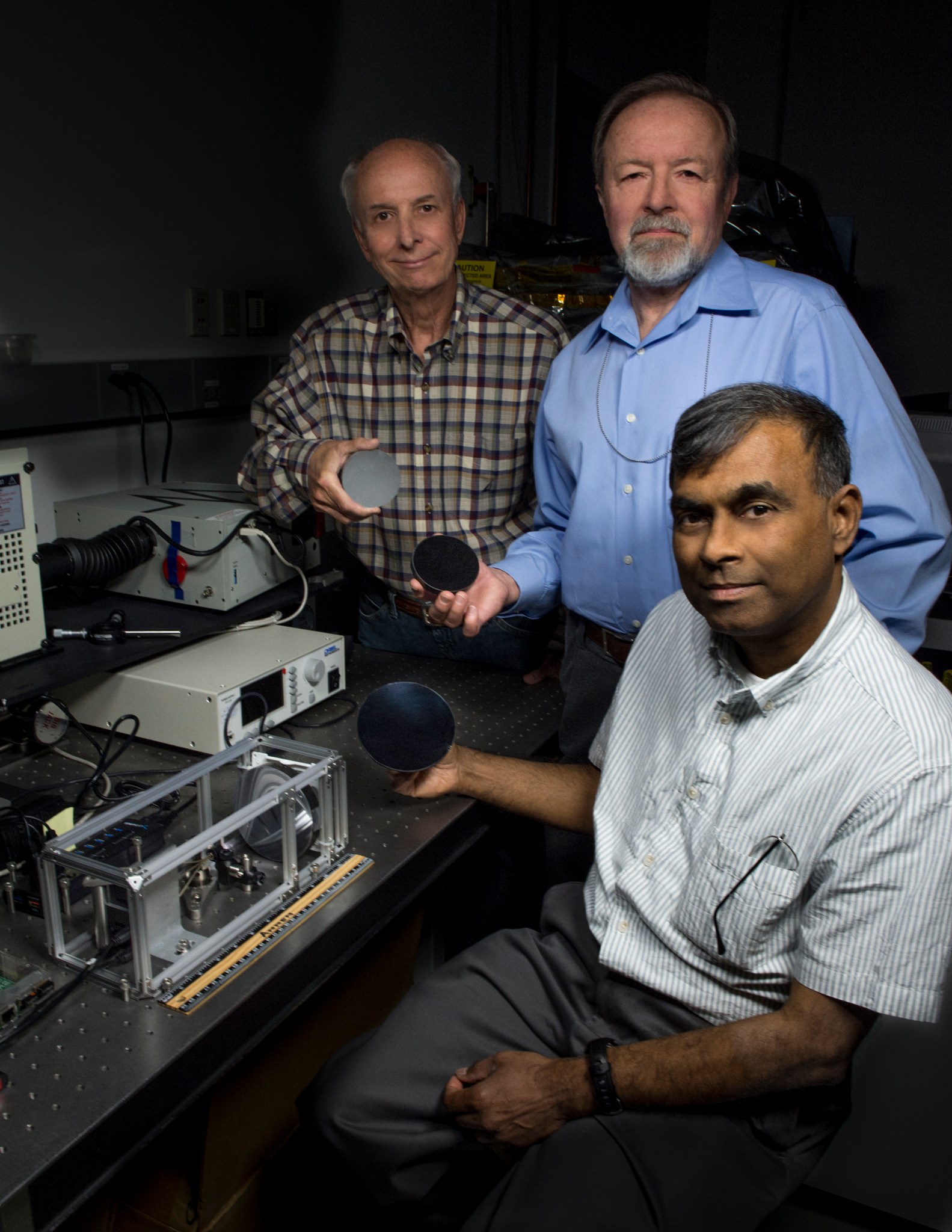 Two men stand and one sits around a table with computers and other engineering tools. They are each holding a small, dark-colored disk.