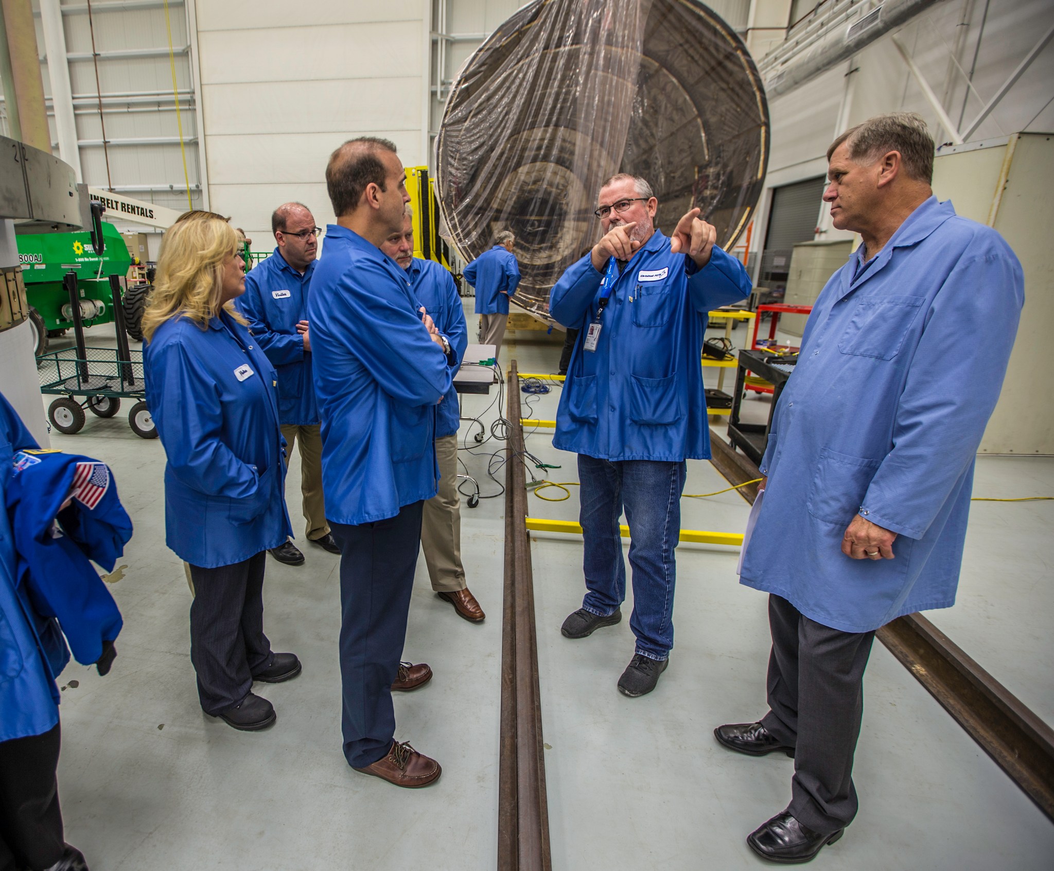 A group of people in blue static jackets stands in a clean room. In the center, one of the people is point off camera while the others listen. Behind them is a large, circular inside of a large rocket covered in plastic.