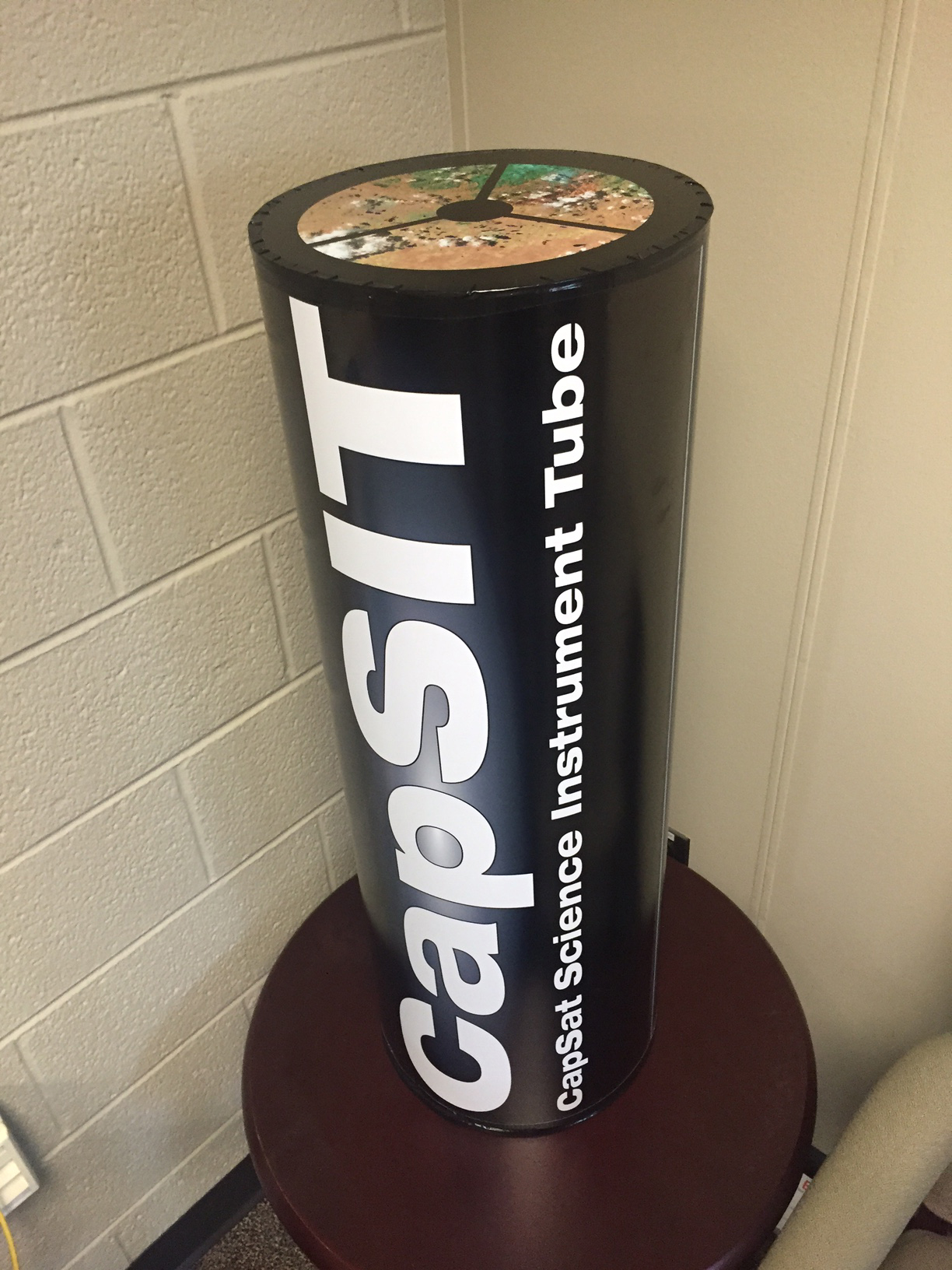 A black cylinder sitting on a brown table. White letters on the side of the cylinder say CapSIT, CapSat Science Instrument Tube.