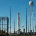 Photo of a slender white rocket on a launch pad at Wallops Flight Facility. The rocket has an American flag and Cygnus and Orbital ATK logos on its conical nose section, and "Antares" written vertically down its side. A tall water tower stands to the rocket's right, and a cagelike structure to its left. Other tanks and fences are visible in the foreground. The sky is clear and bright blue.