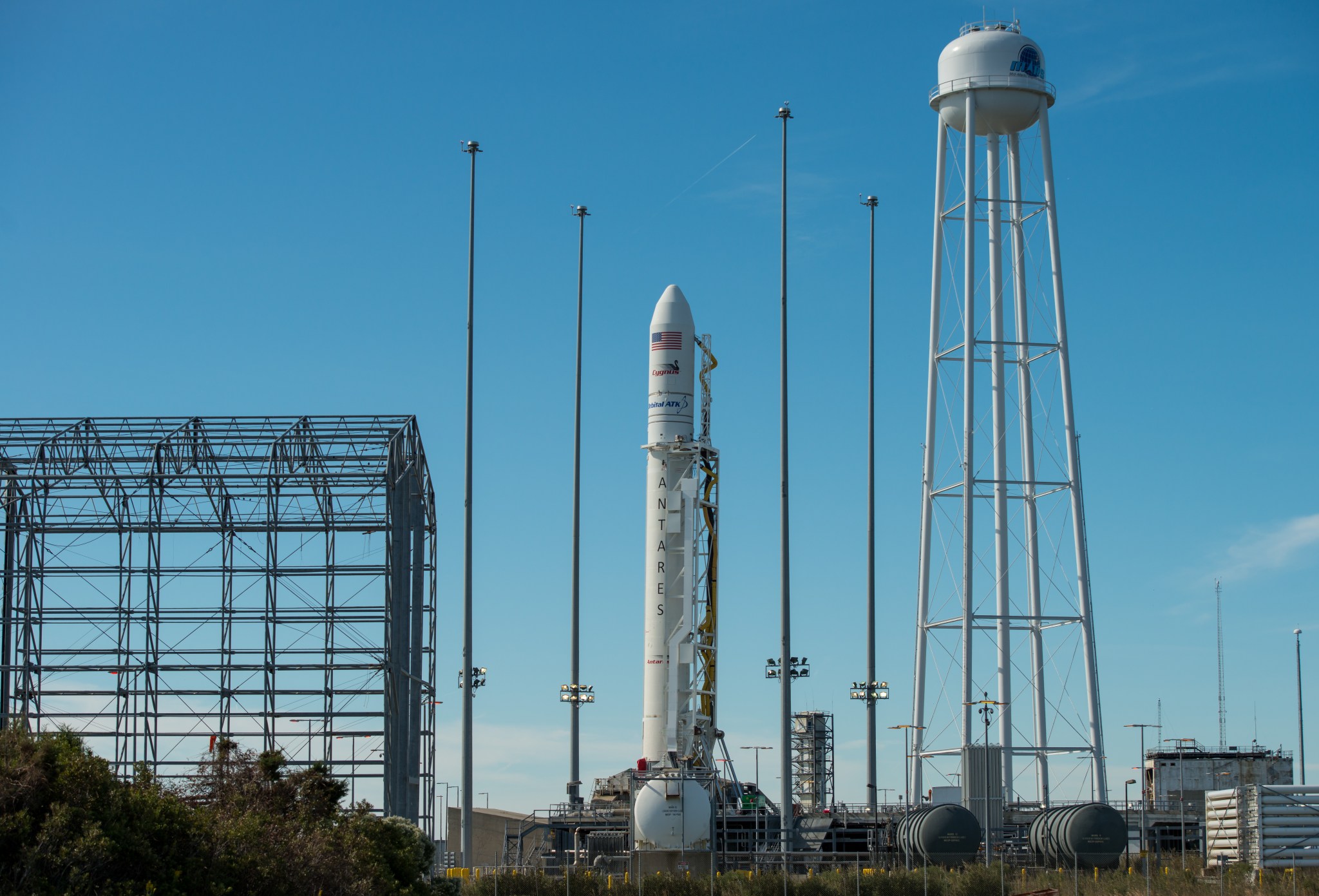 Photo of a slender white rocket on a launch pad at Wallops Flight Facility. The rocket has an American flag and Cygnus and Orbital ATK logos on its conical nose section, and "Antares" written vertically down its side. A tall water tower stands to the rocket's right, and a cagelike structure to its left. Other tanks and fences are visible in the foreground. The sky is clear and bright blue.