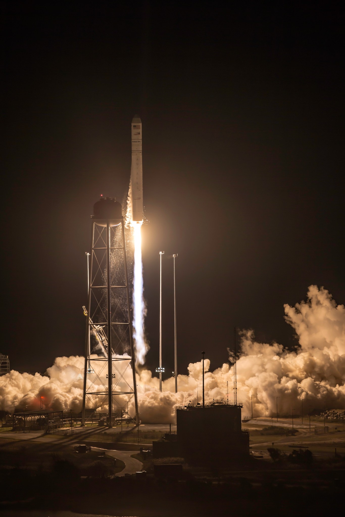 A nighttime shot of a white rocket just seconds launching off the launch pad, flanked by two bright white lights, with a plume a smoke just underneath. The buildings and ground around the rocket are softly lit by red lights near the ground, and the whole image is soft from hazy humidity.