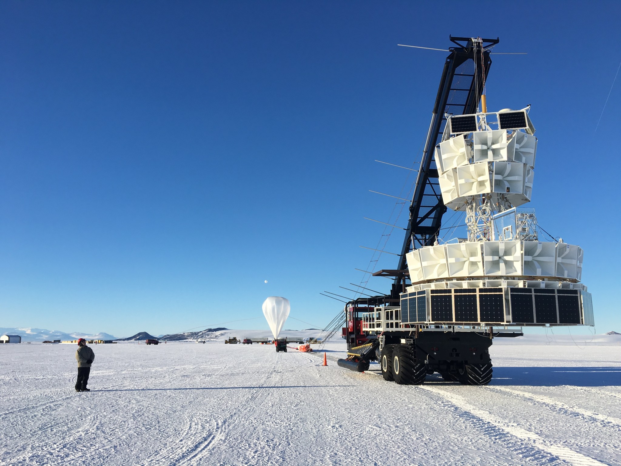 The ANITA payload, an instrument made up of many white boxes formed a circle  tube structure, hangs from a crane with a scientific balloon in the background against a blue sky and white, snowy surface.