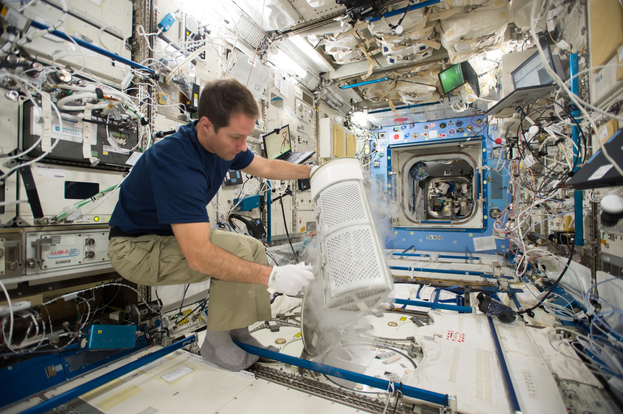 ESA astronaut Thomas Pesquet, wearing a short sleeved blue shirt, khaki pants, grey socks, and white gloves, holds a compartment from the station’s freezer. The white compartment is wedge-shaped with a screen-like surface. Water vapor rises from it like smoke.