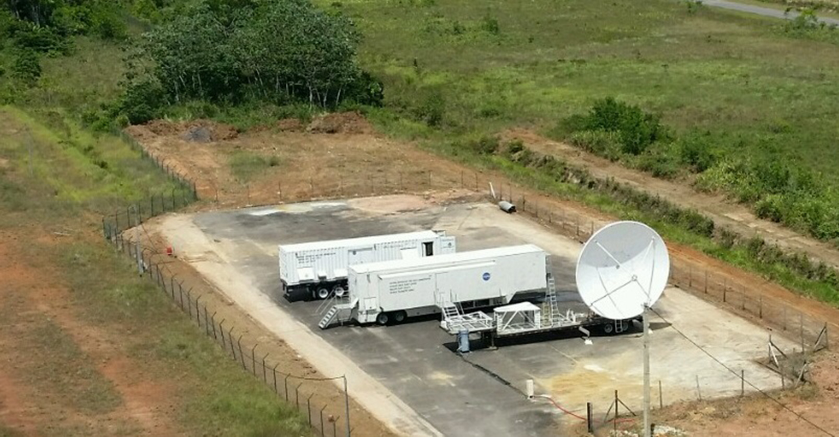 Two white trailers and a large white antenna are setup on an asphalt pad in the middle of a field.