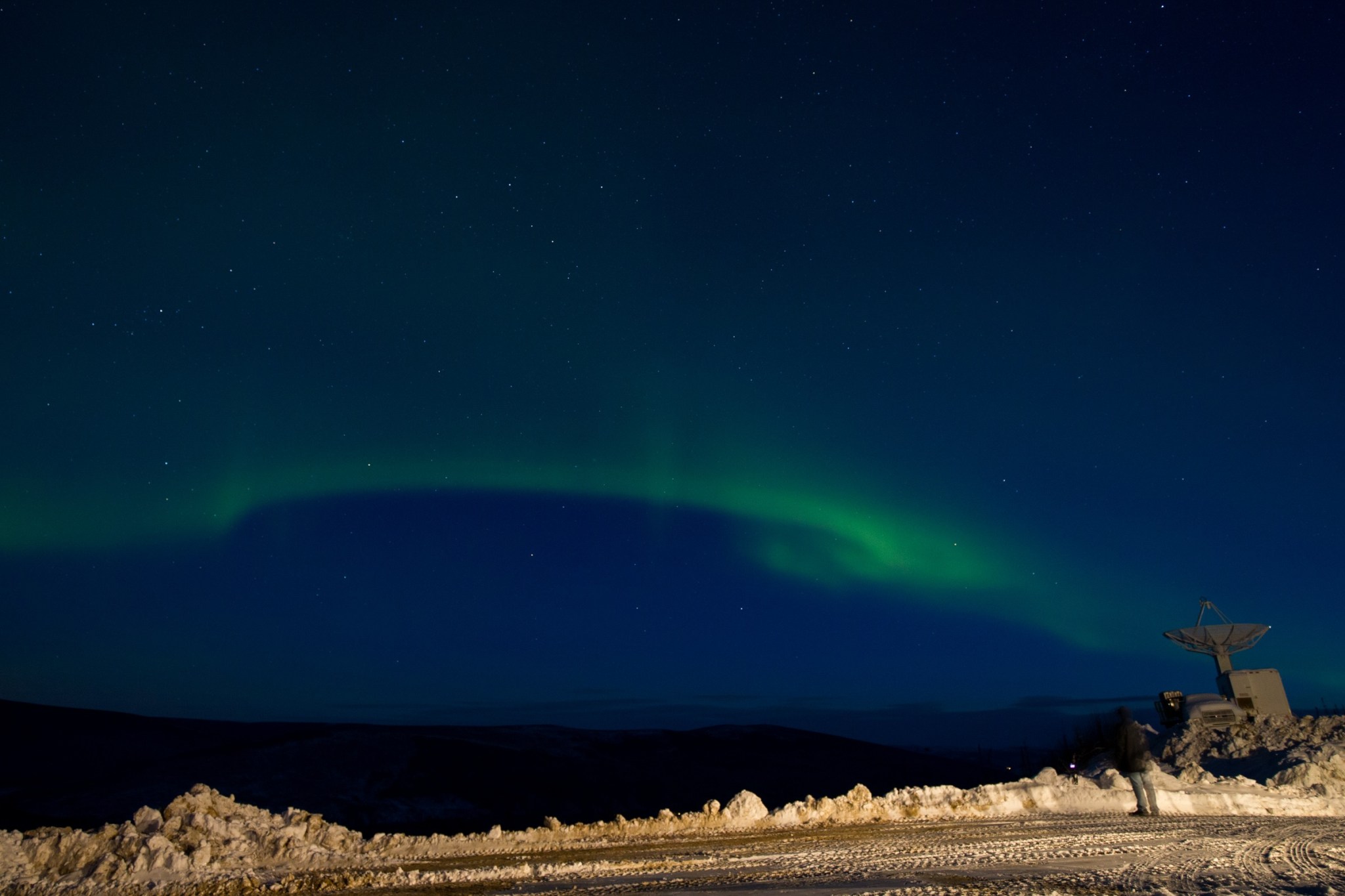 A thin green streak of aurora move across a dark blue sky, with a blanket of snow in the foreground.