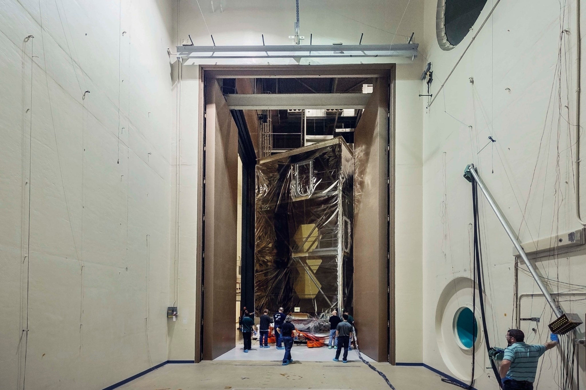 Another view of the transparent box from inside the acoustic chamber. Tall beige walls make up the sides, and huge heavy doors are open at the far end, where the telescope in its box is being moved.