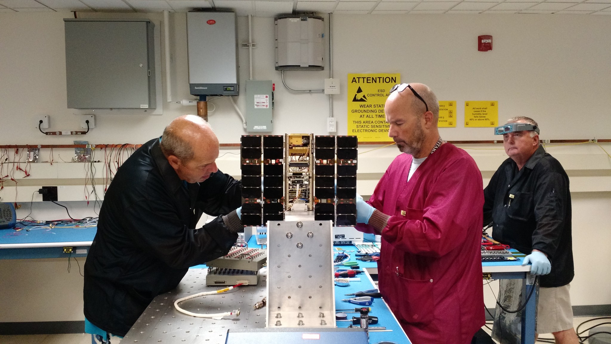 Two people work on the IceCube smallsat, a small open rectangular structure with solar panels on each side. A third person looks on from the background.