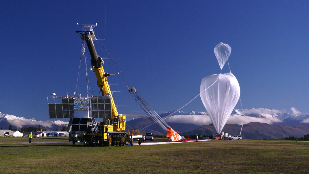 A scientific balloon is to the right, and appears as a plastic, upside down teardrop. A tube attached to the top of the balloon leads down to the ground. A crane to the left holds a large payload structure with many solar panels. In the background is a small treeline and a mountain range with a dusting of snow ontop.