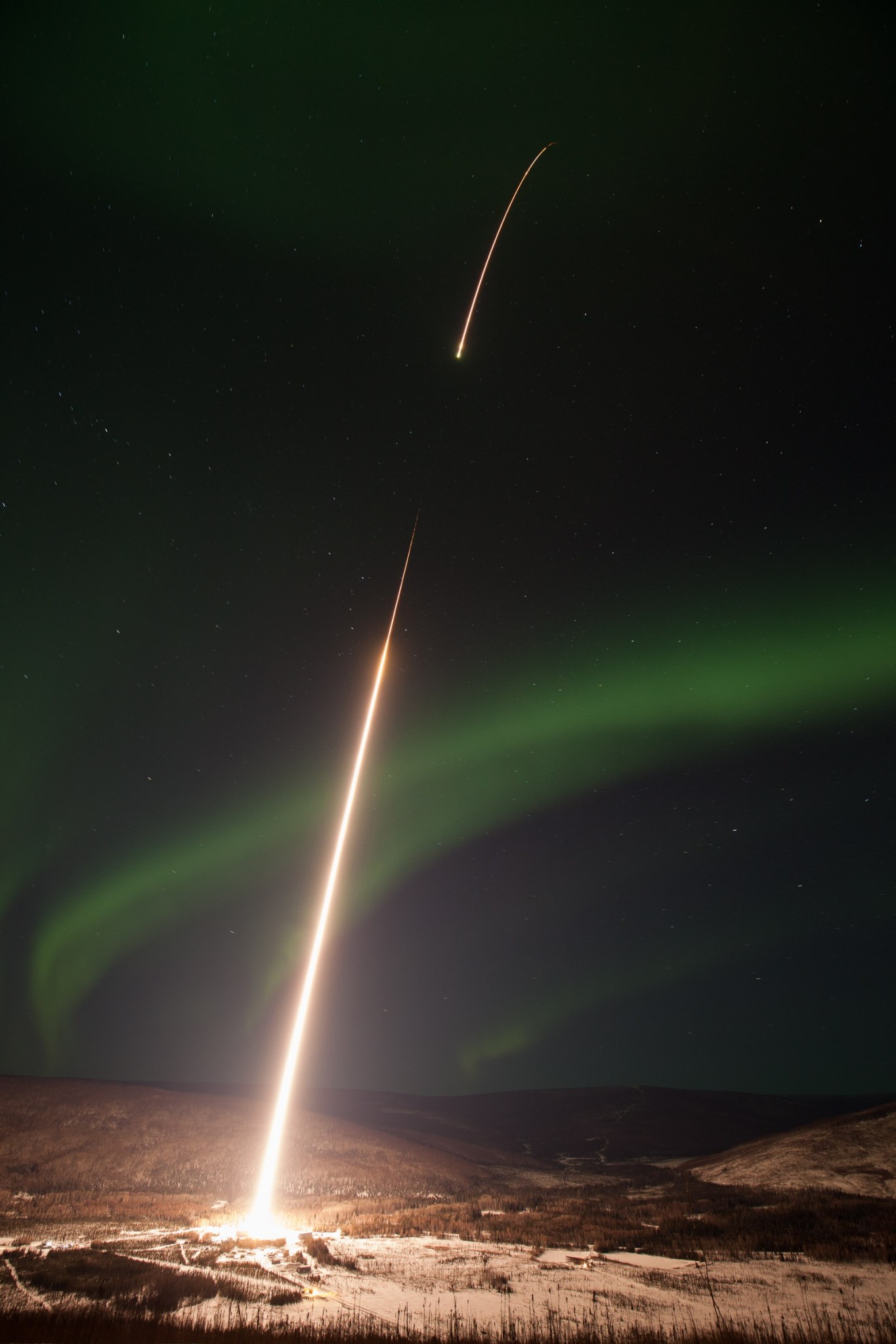 A long-exposure photograph of a souding rocket launch, represented by a white streak broken in the middle. A soft green aurora is in the background against a snowy landscape.
