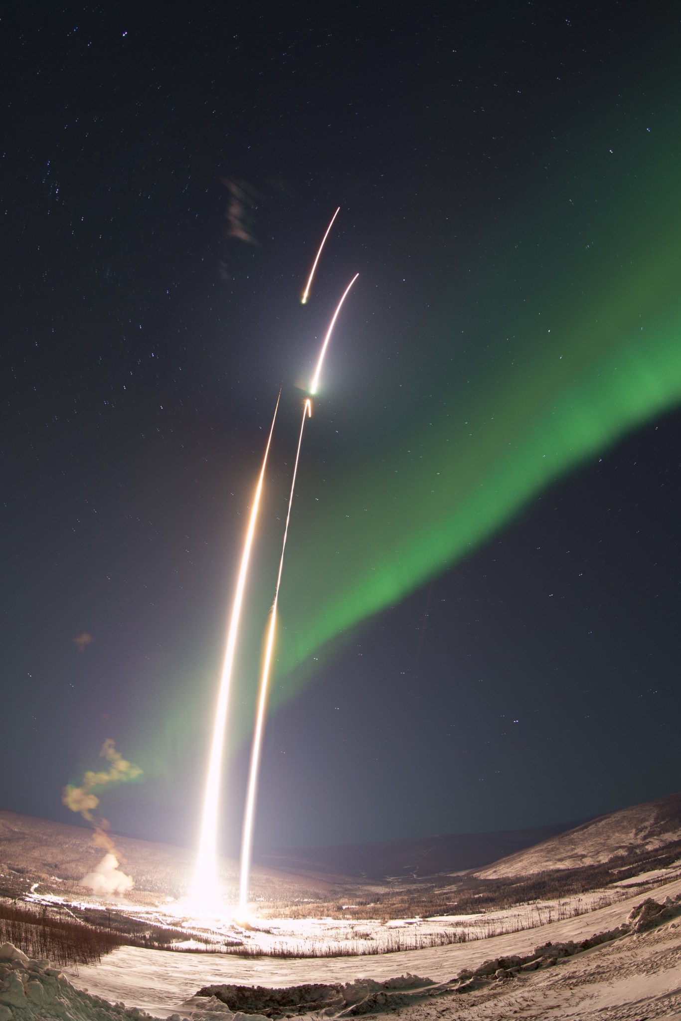 A composite, long-exposure image of two sounding rockets launching, represented by two white streaks. A bright green aurora is in the background against a snowy landscape.