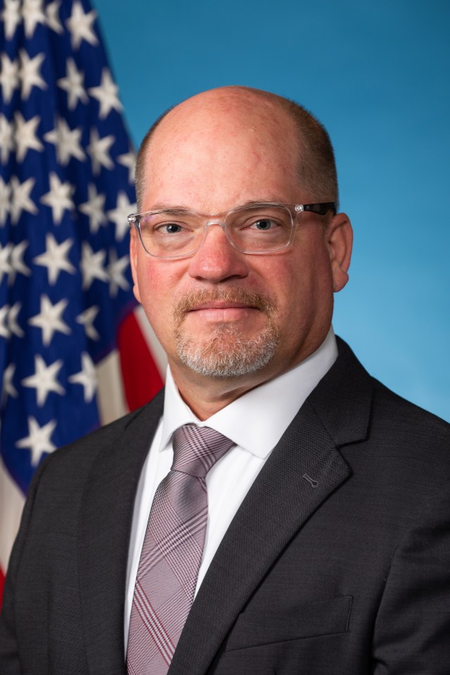 Chad Brown in glasses wearing a suit and tie poses for a headshot photograph in front of an American flag and blue backdrop.