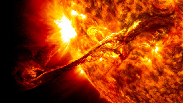 An image of a filament, a long, slender eruption snaking away from the Sun's surface. The Sun appears as a huge, glowing red-orange ball with a swirling surface, filling the right side of the image.