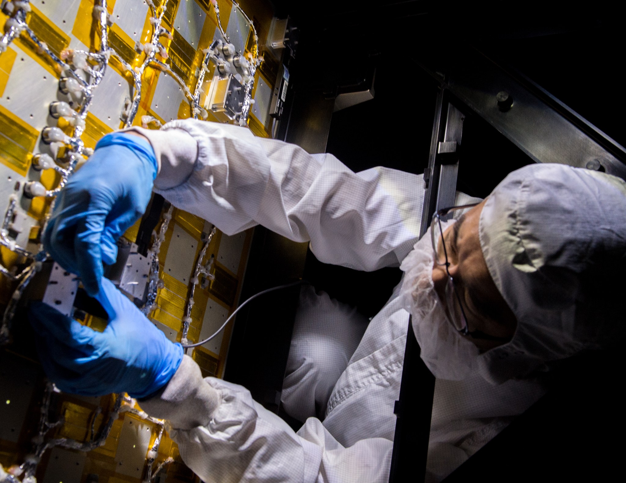 A person wearing a cleanroom protective gear, including a hat, facemask, body suit, and gloves, leans over an electronis panel. The panel has gold and white rectangles with silver-wrapped wires running between the rectangles.