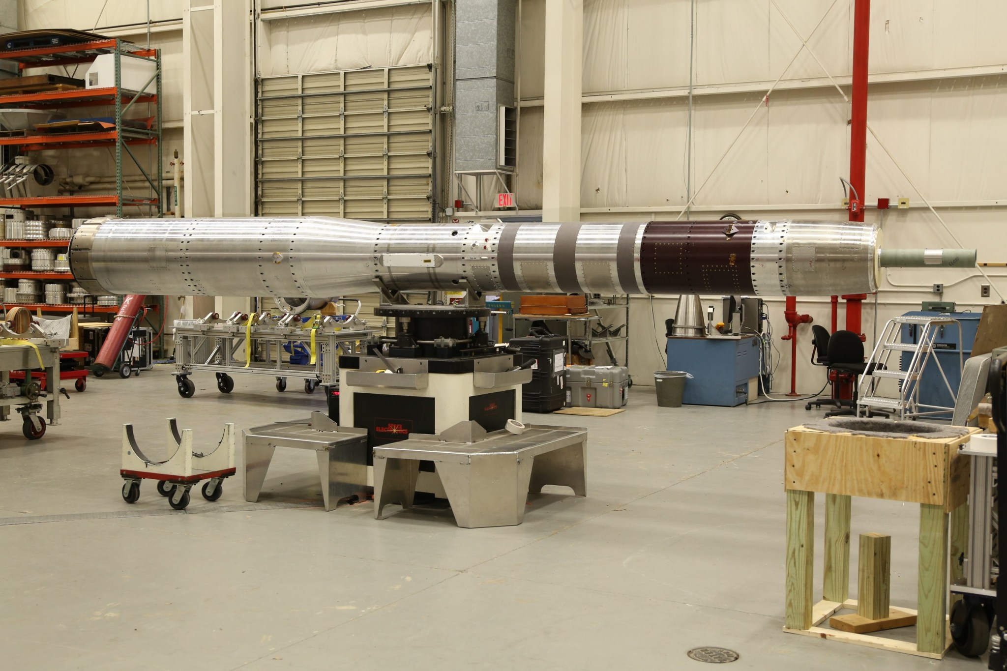 A long, metal payload section of a sounding rocket is set on a metal stand inside a large clean room with other various pieces of sounding rocket parts around.