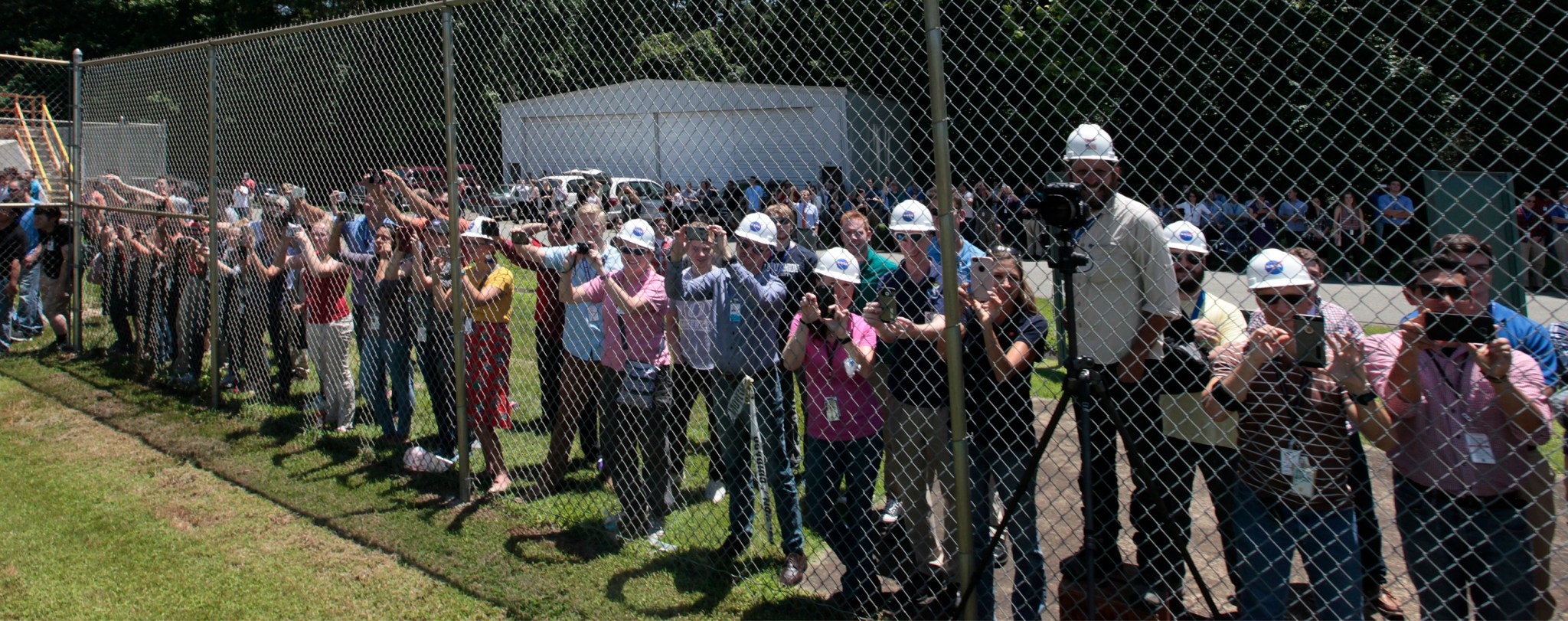 Many people in hard hats stand against a chain-link fence with cameras.