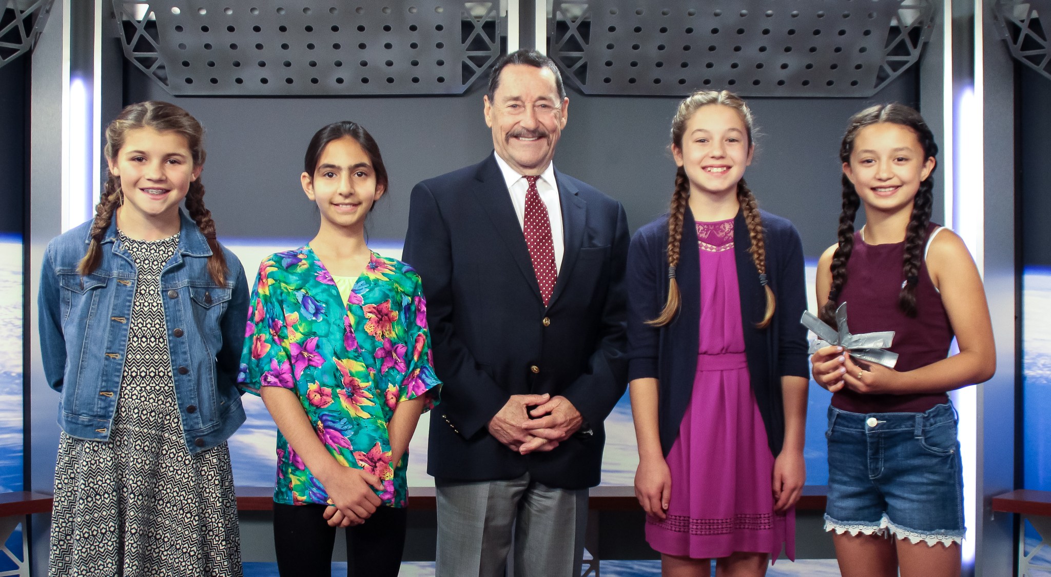 Winners of OPSPARC challenge pose with Peter Cullen. The winners are four young girls standing in a line with Peter in front of a backdrop that looks like space. 