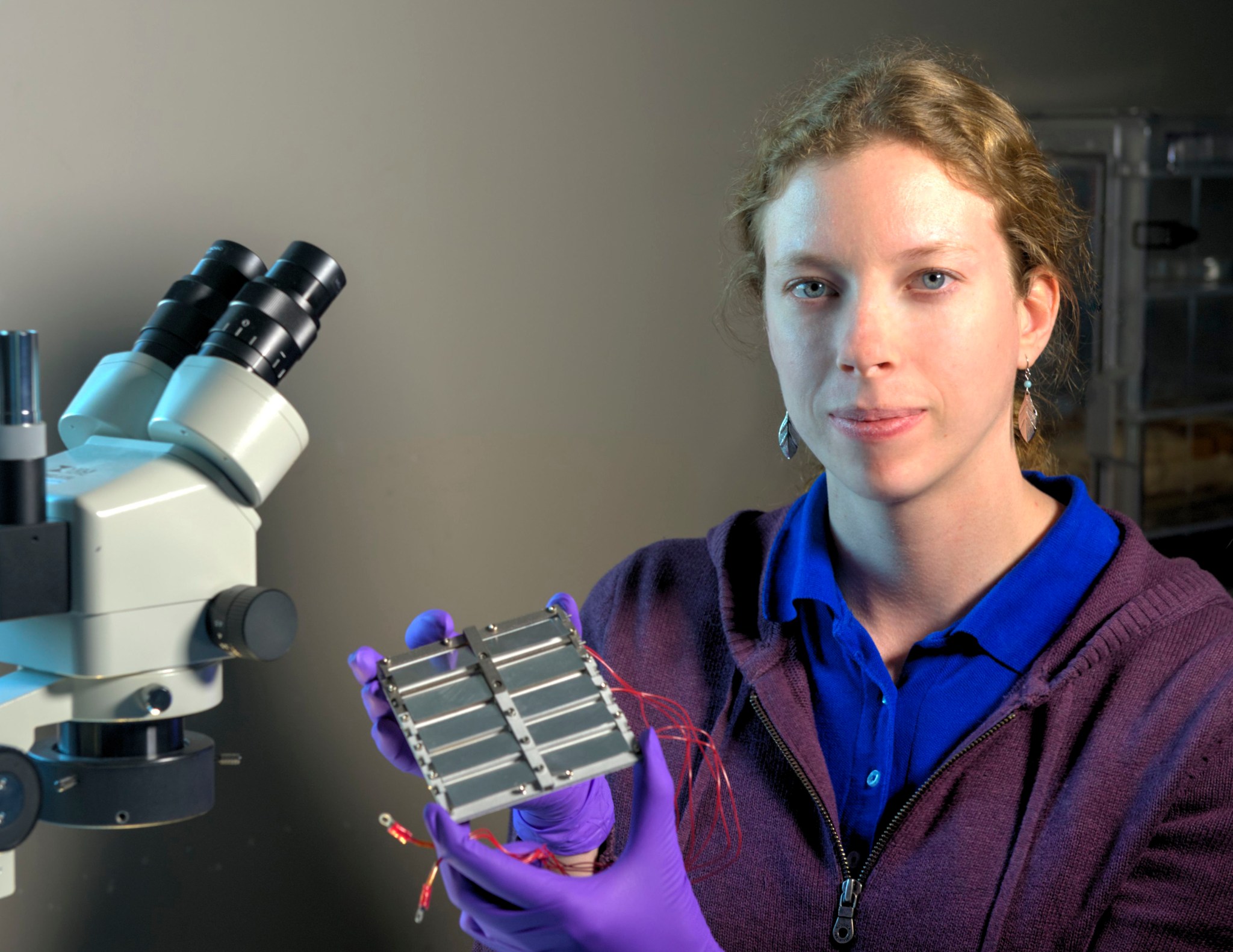 Allison Evans, woman with fair skin and light hair holds technology with red wires next to a microscope