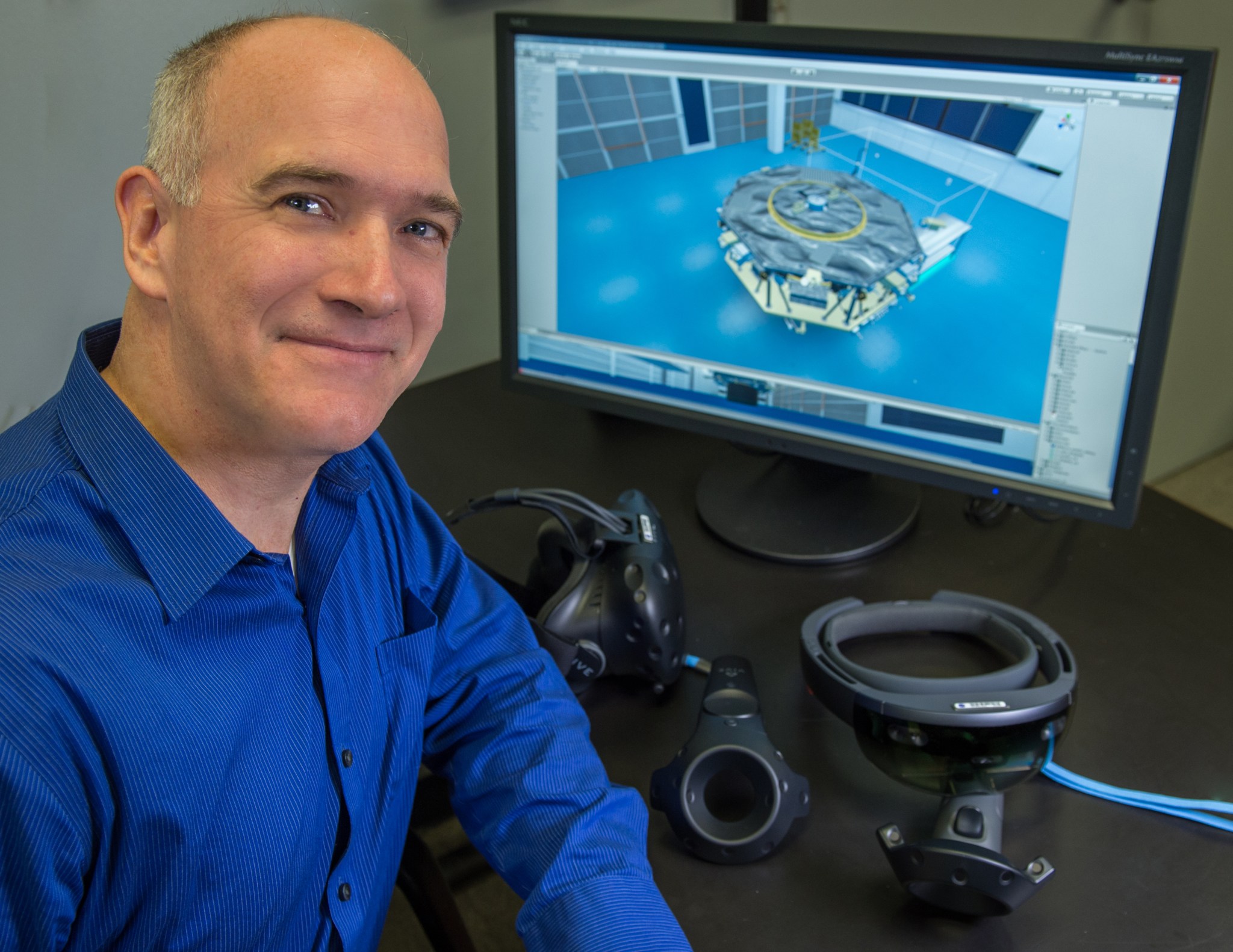 Man with fair skin wearing a blue dress shirt smiles next to a computer monitor showing VR/AR