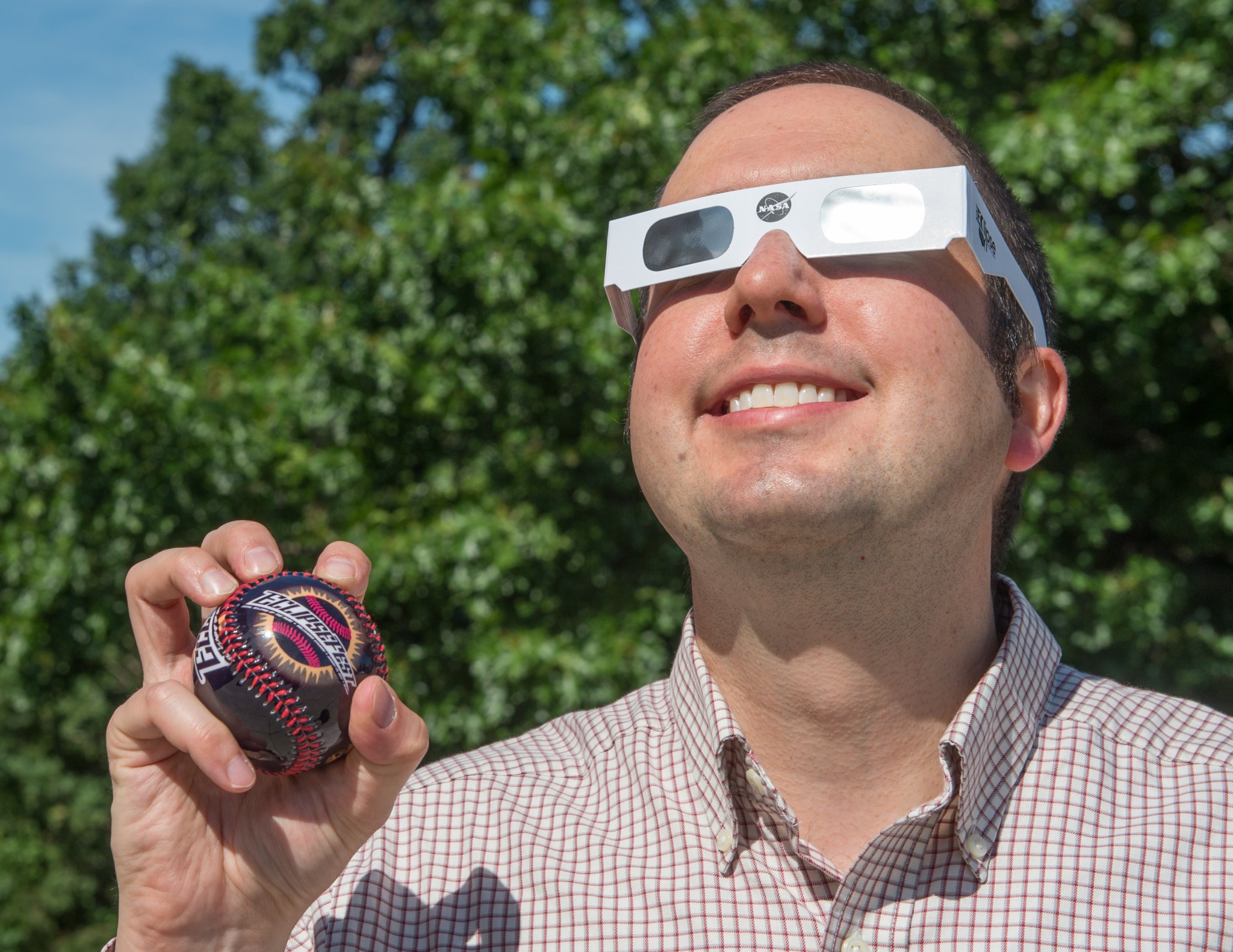 Noah Petro looking up at the Sun, wearing dark eclipse glasses and holding a baseball that says Eclipsefest.