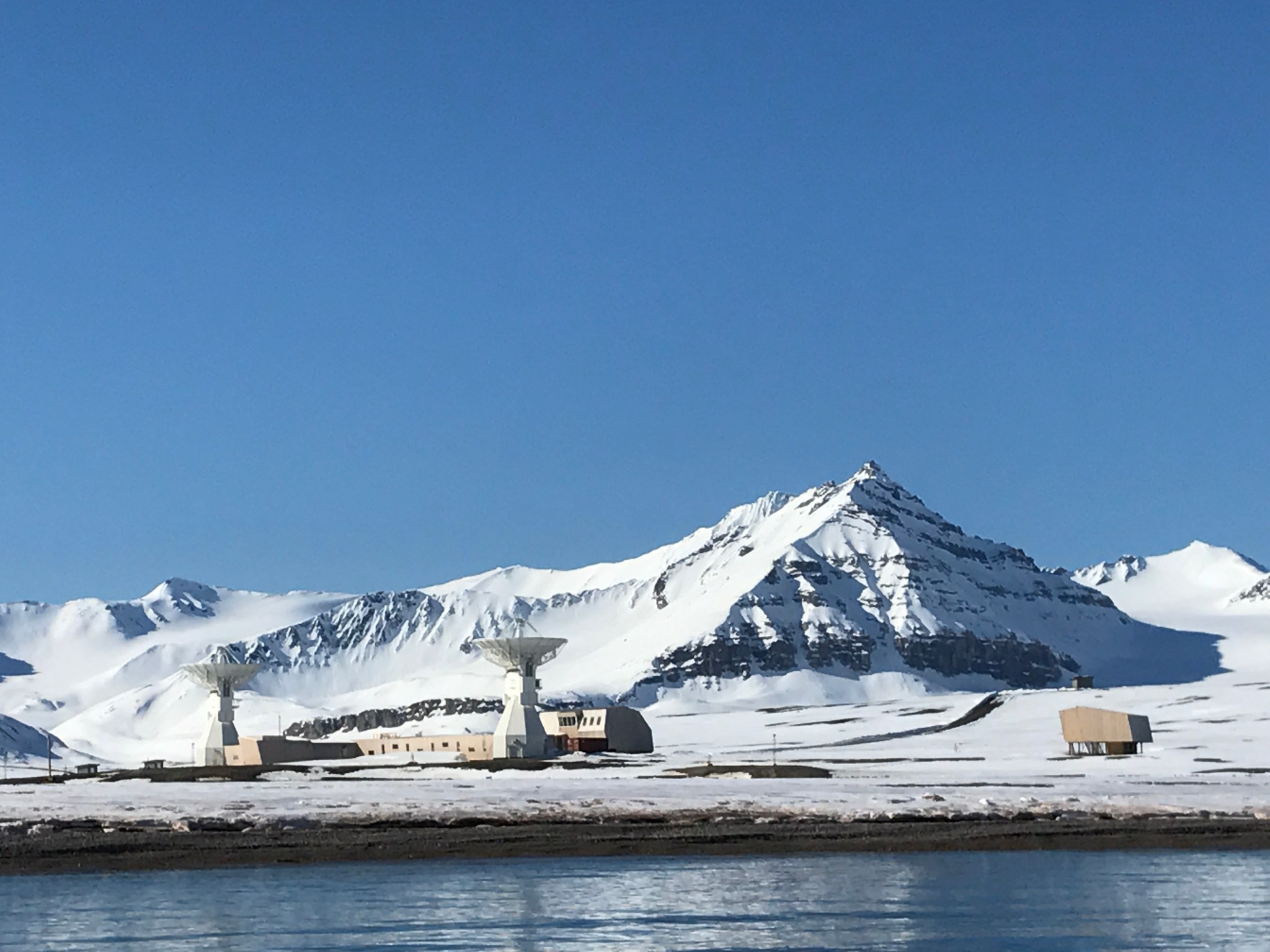 The scientific base of Ny-Ålesund, Svalbard, as seen from the sea.  The base is made of small tan buildings with two white satellite dishes pointed to the sky. Snow covered mountains rise just behind the base.
