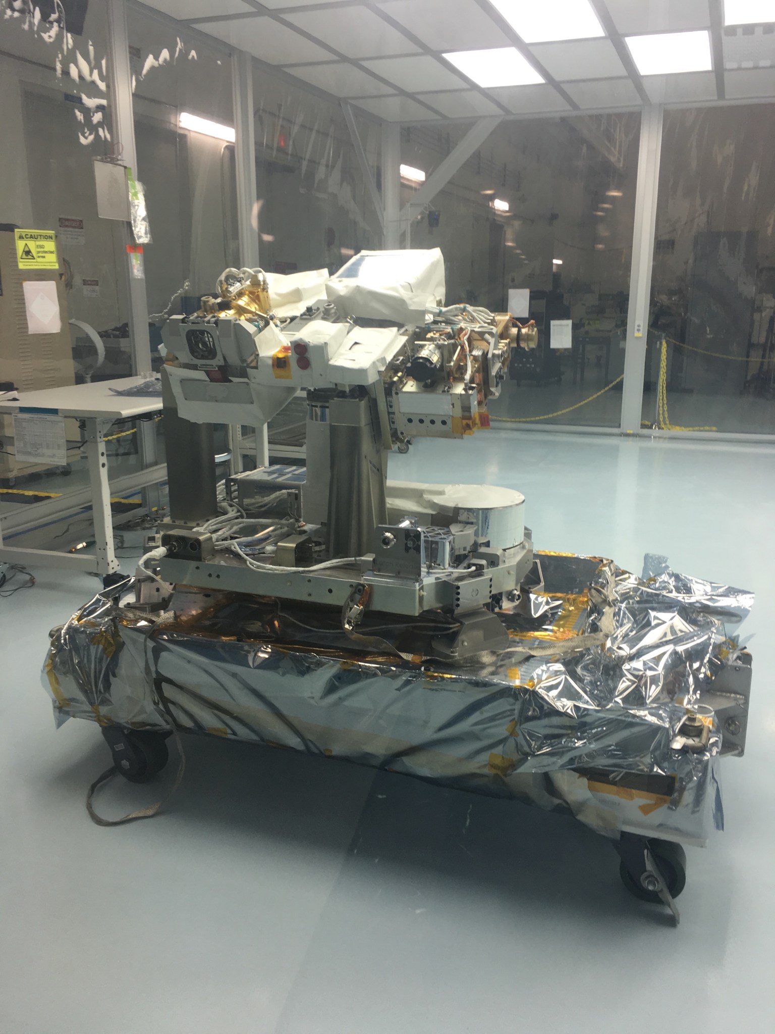 TSIS-1 in the clean room. The spacecraft comprises several meal rectangular prisms on a thin metal neck. Components are wrapped in silver foil with gold tape. It sits on a low, flat wheeled cart.