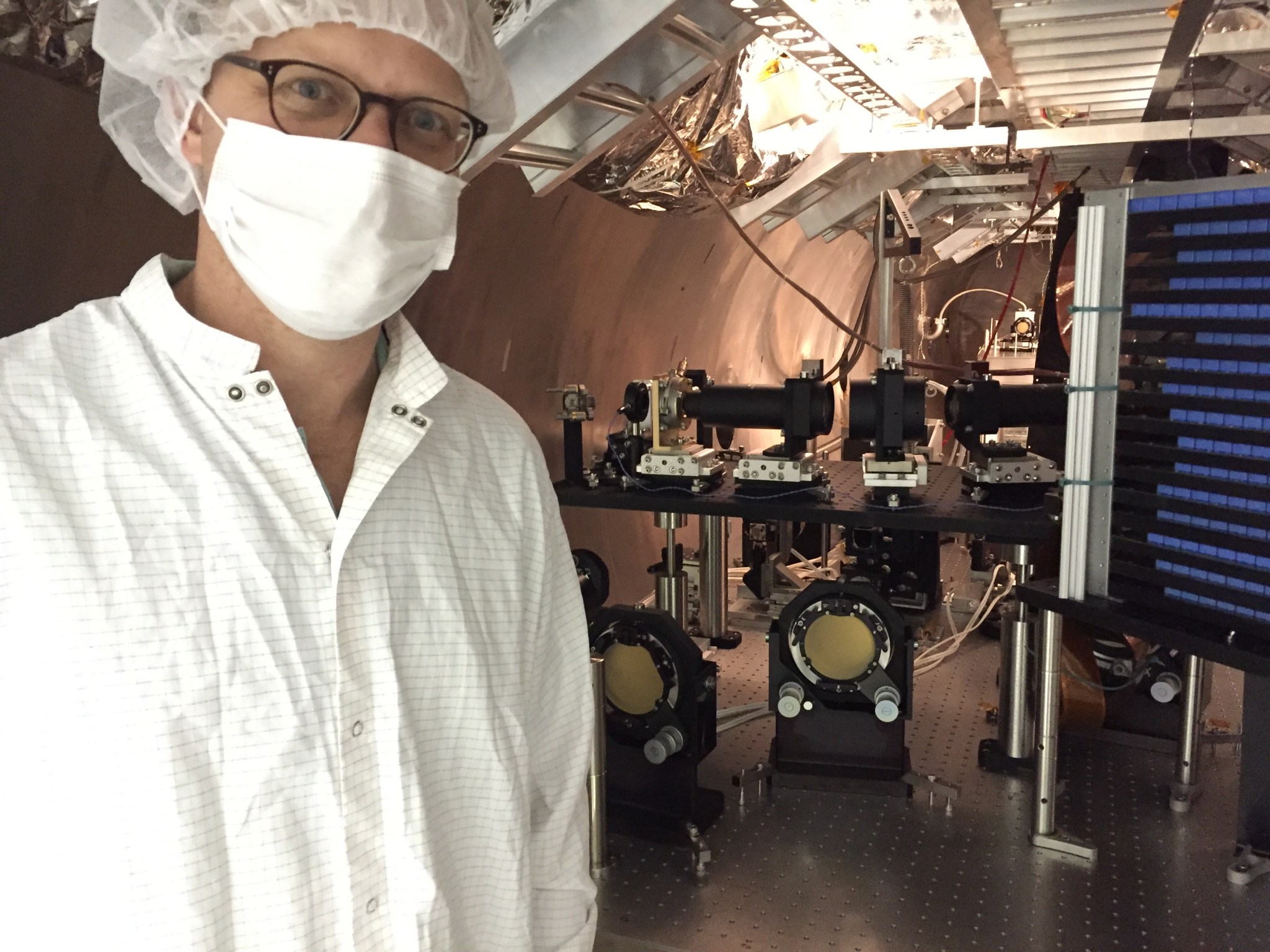 Instrument Scientist Michael McElwain wears a lab coat, mask and hair net, standing in the High-Contrast Imaging Testbed at NASA’s Jet Propulsion Laboratory 