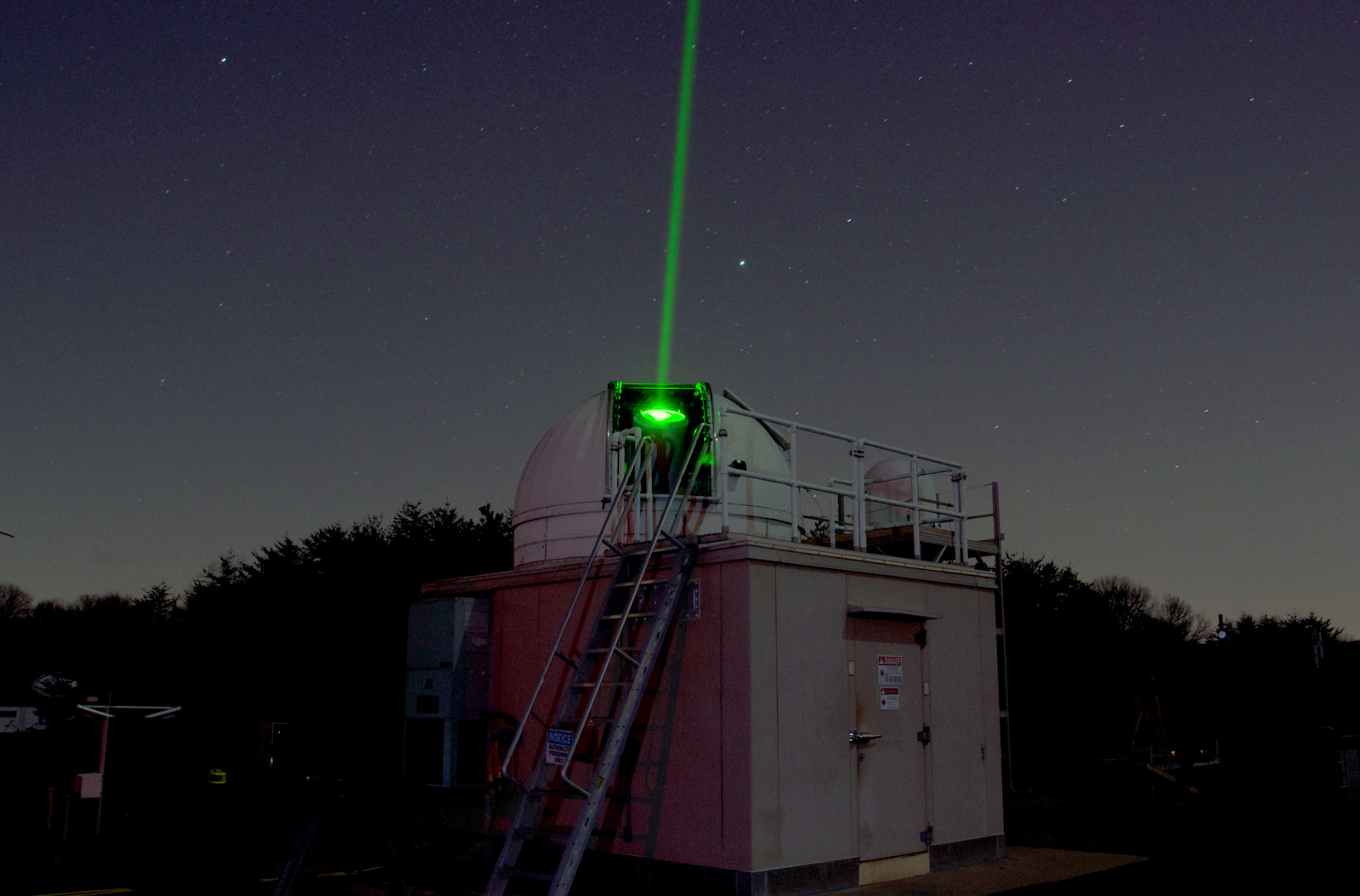 The Next Generation Satellite Laser Ranging station, which has a white metal dome, with a small segment open. A green laser beam shoots out of the dome opening toward the night sky.