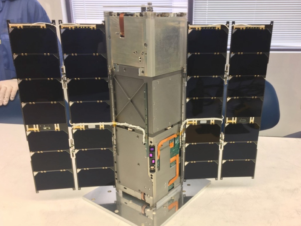 The RAVAN small satellite sitting on a table. It's a long silver rectangular prism, sitting up on one of the small sides. Solar panels jut out from each side like wings.