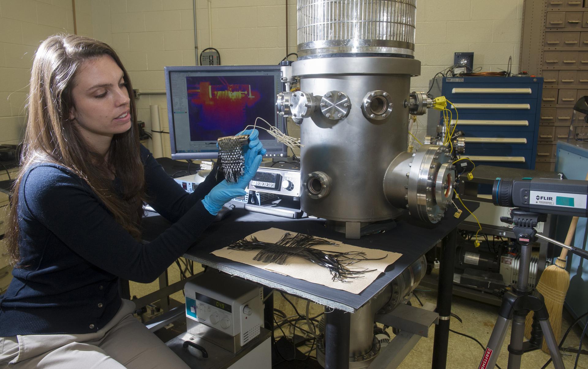 A women in a blue shirt and gloves holds an object in front of a thermal camera, with the cameras output on a screen behind her.