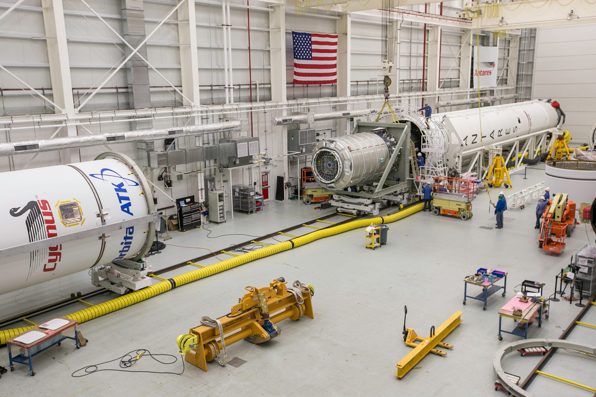Inside a large clean room with the Antares rocket on its side. The rocket is open, with the top portion off on the left side of the image. The Cygnus payload is attached to the bottom half of the rocket, moved away from the top of the rocket. The rocket is white with the words "Antares" running down the side.