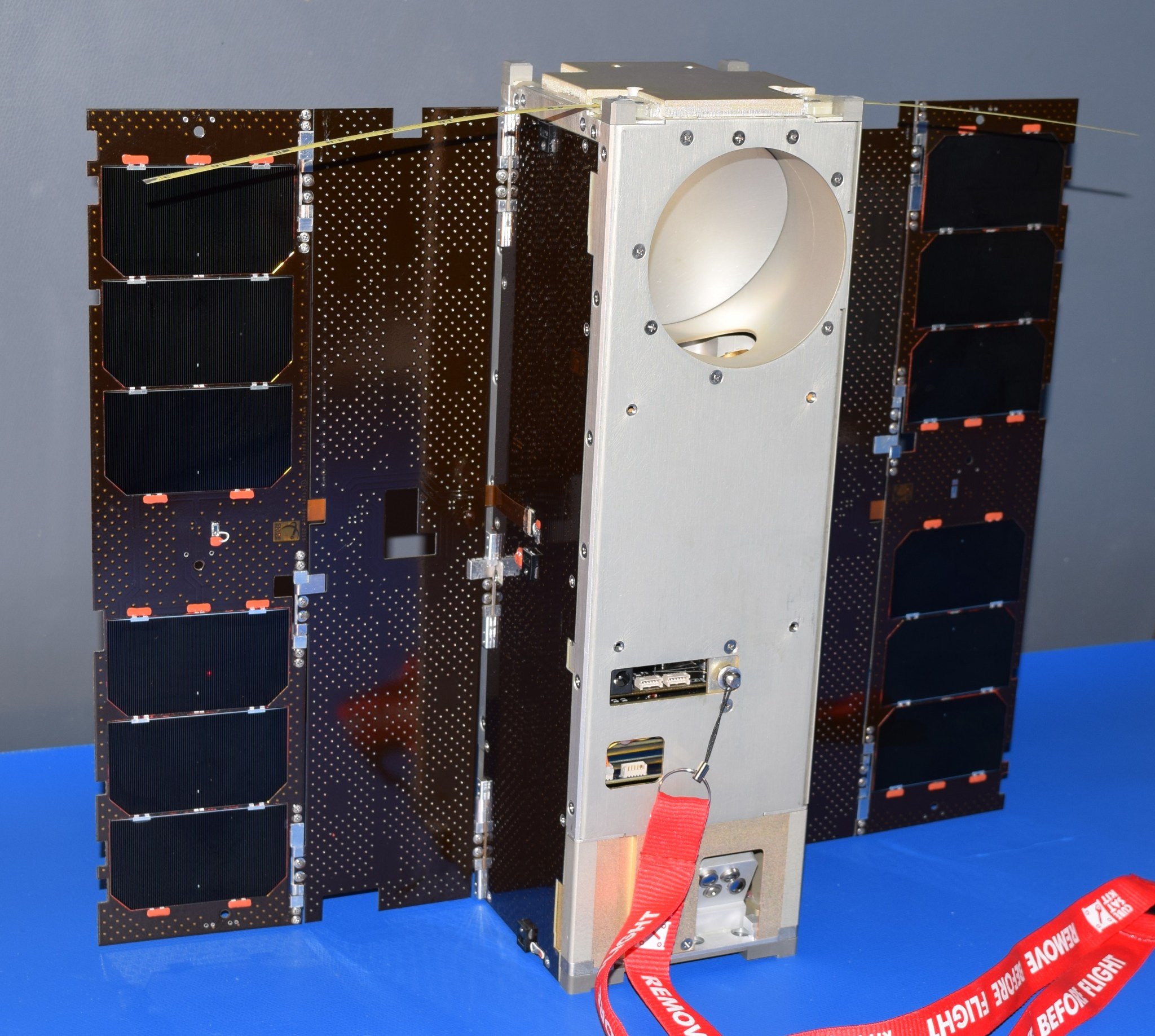A small satellite sitting on a blue table. The satellite is a long, thin rectangular prism with a solar panel on either side like wings. A red lanyard that says "Remove Before Flight" is hooked to the belly of the satellite.
