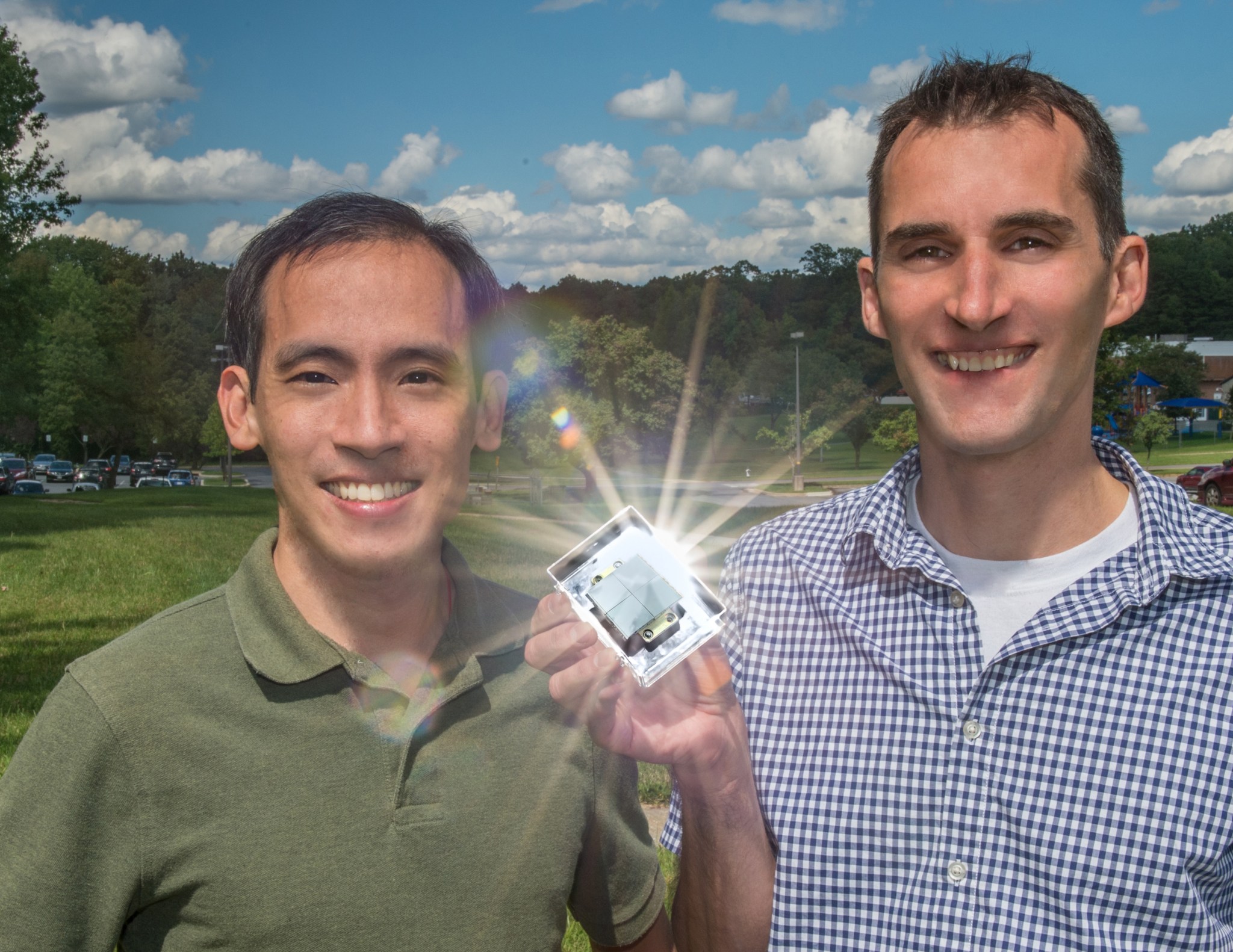 Two men smiling at the camera. One of them is holding a small metal box that is reflecting a huge beam of light in several directions.