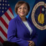 A black female in a blue jacket poses for a picture in front of the American flag and the NASA flag