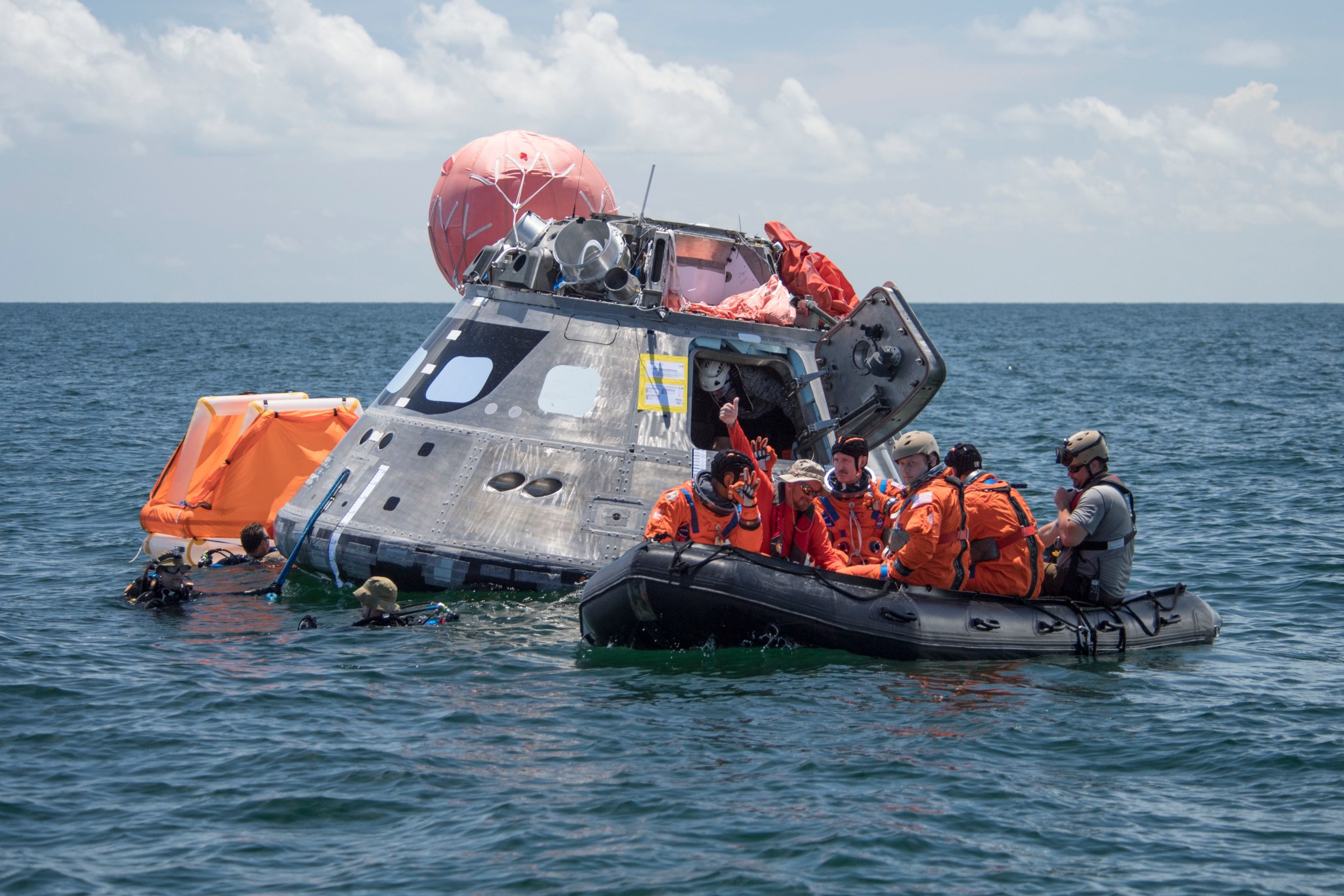 Orion capsule floats in sea with crew in orange flight suits in raft nearby