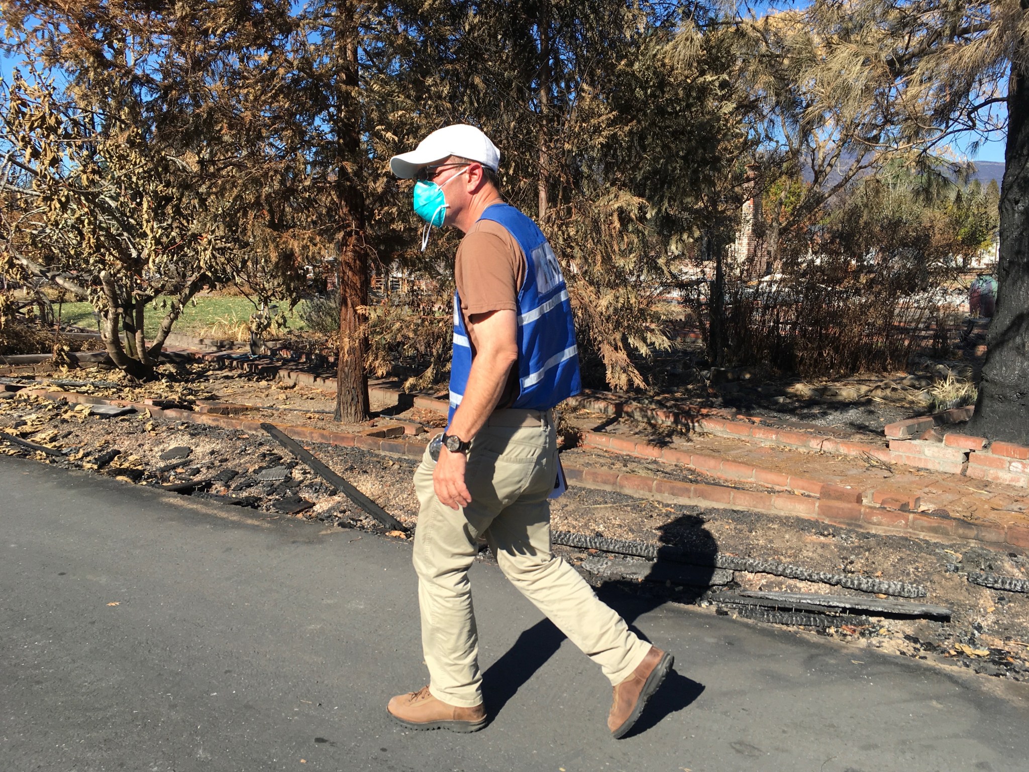 A man is shown wearing a blue vest, brown t-shirt, khaki pants, baseball cap, and a surgical mask. Behind him, burned trees and shrubs can be seen as he walks along the road. 