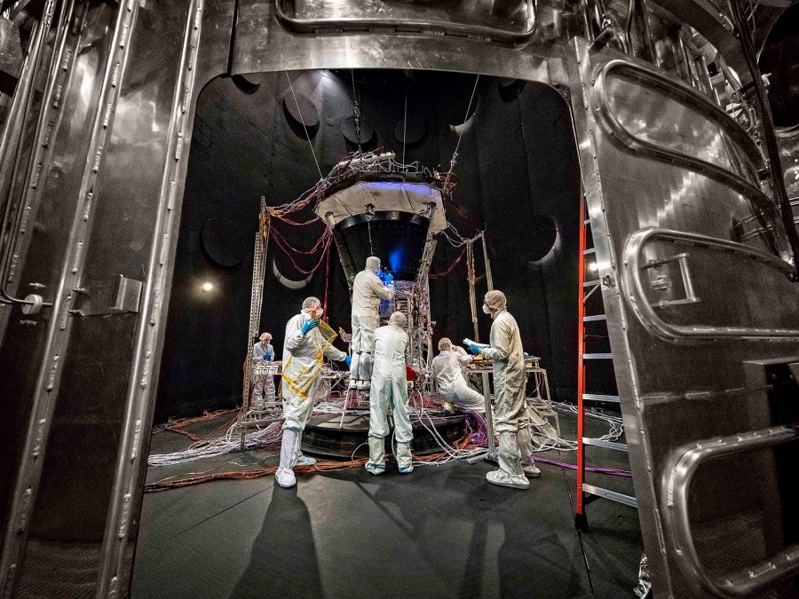 People in clean suits work on the spacecraft inside a large chamber