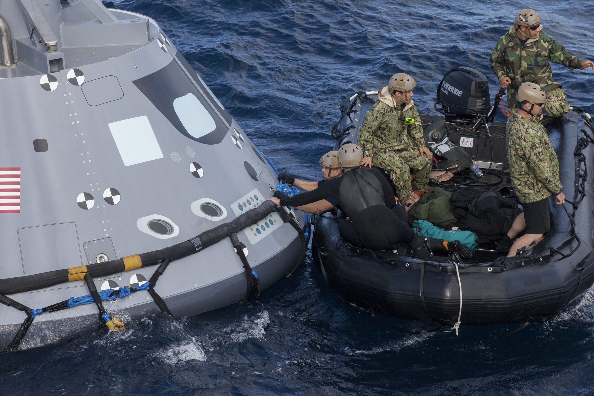 U.S. Navy divers and other personnel in a Zodiac boat secure a harness around a test version of the Orion crew module