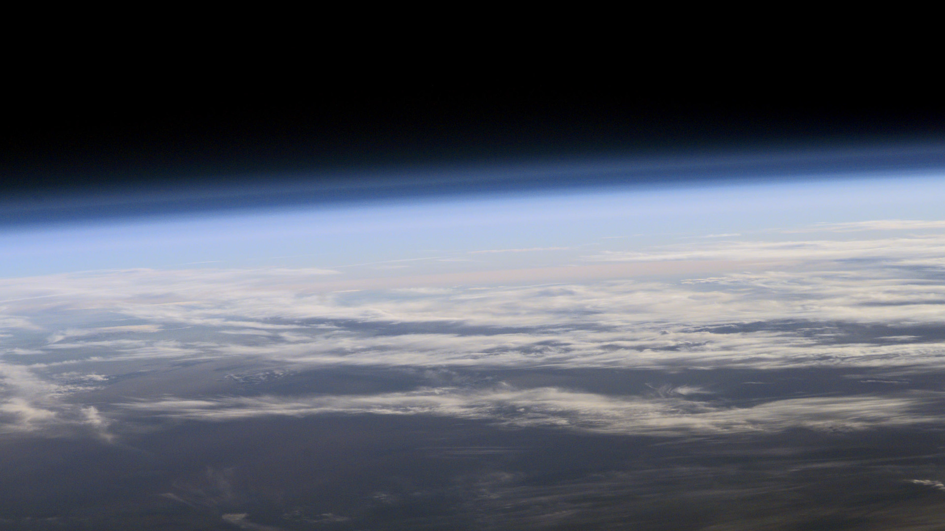 A view from space of Earth's atmosphere, looking out across the upper layer of the atmosphere into space. Faint, partly transparent blue and pink gradients are visible above the cloud line.