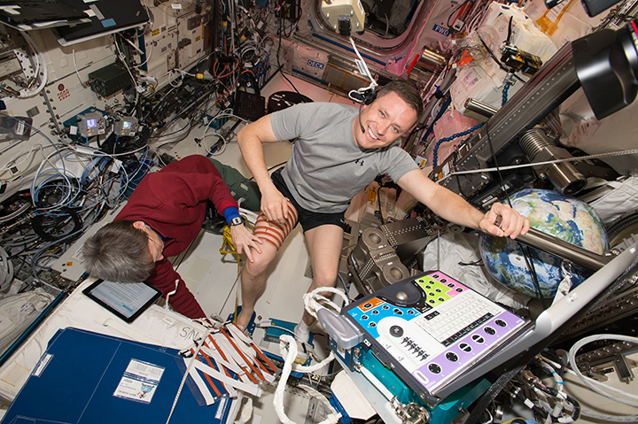 Fisher is wearing a gray shirt, black shorts, and white socks, with his feet tucked into straps, and is holding on to a bar with his left hand. Whitson, wearing a red sweatshirt and green pants, applies strips of red tape to Fisher’s leg that are used to guide an ultrasound of his muscles. There is a multi-colored control panel in front of Fisher and an inflatable Earth ball behind it, and the wall behind them has multiple laptops, equipment, and coiled cords. 