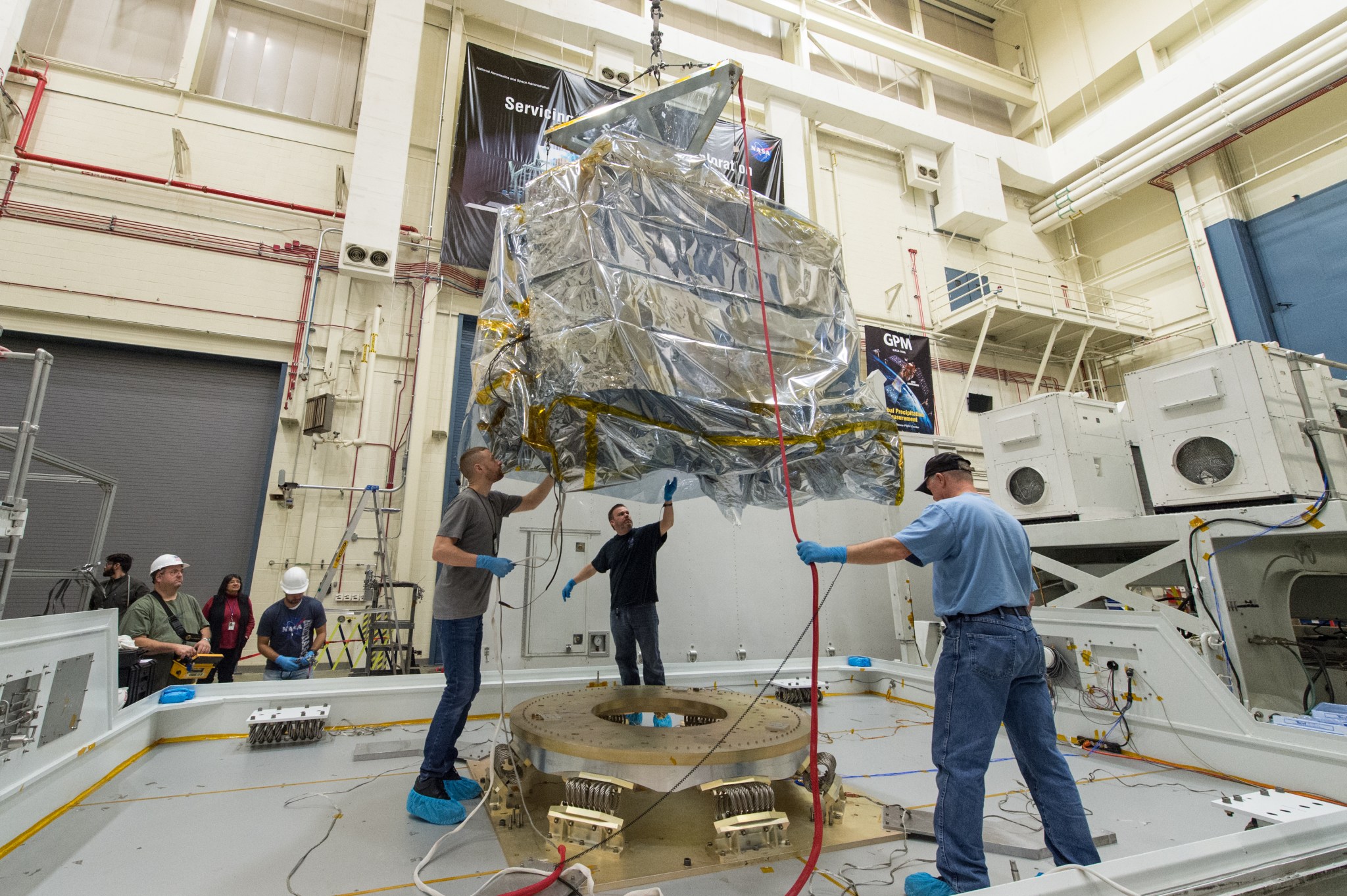The ATLAS team carefully lowers the instrument onto a platform, held up by wires. The spacecraft is wrapped in silver foil.