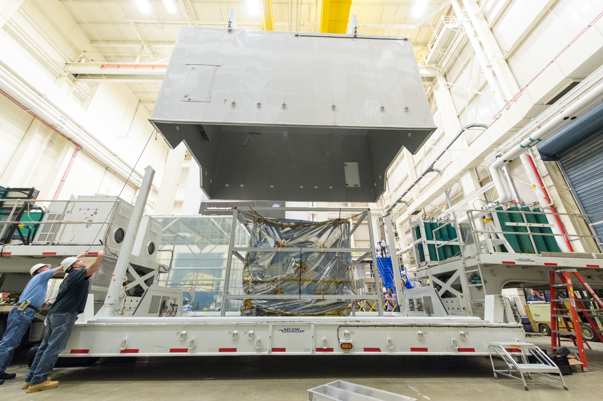 Crews at NASA Goddard lower a cover onto the transporter truck. The spacecraft is encased in silver foil and the lid of the cover is being lowered from the ceiling, like a big box.
