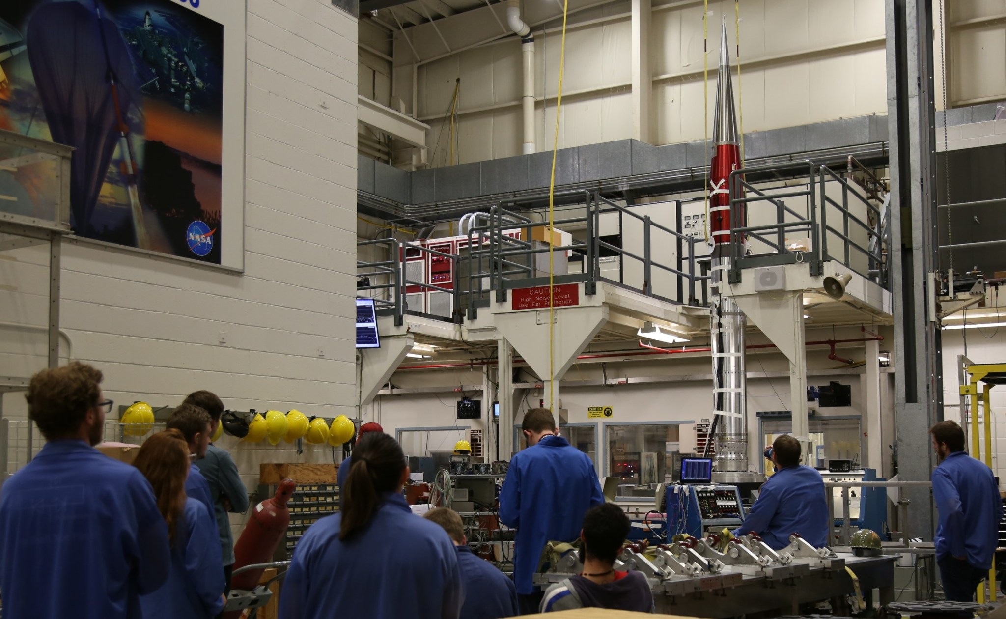 Students in blue anti-static coats watch the USIP sounding rocket payload going through vibration testing vertical on a stand.