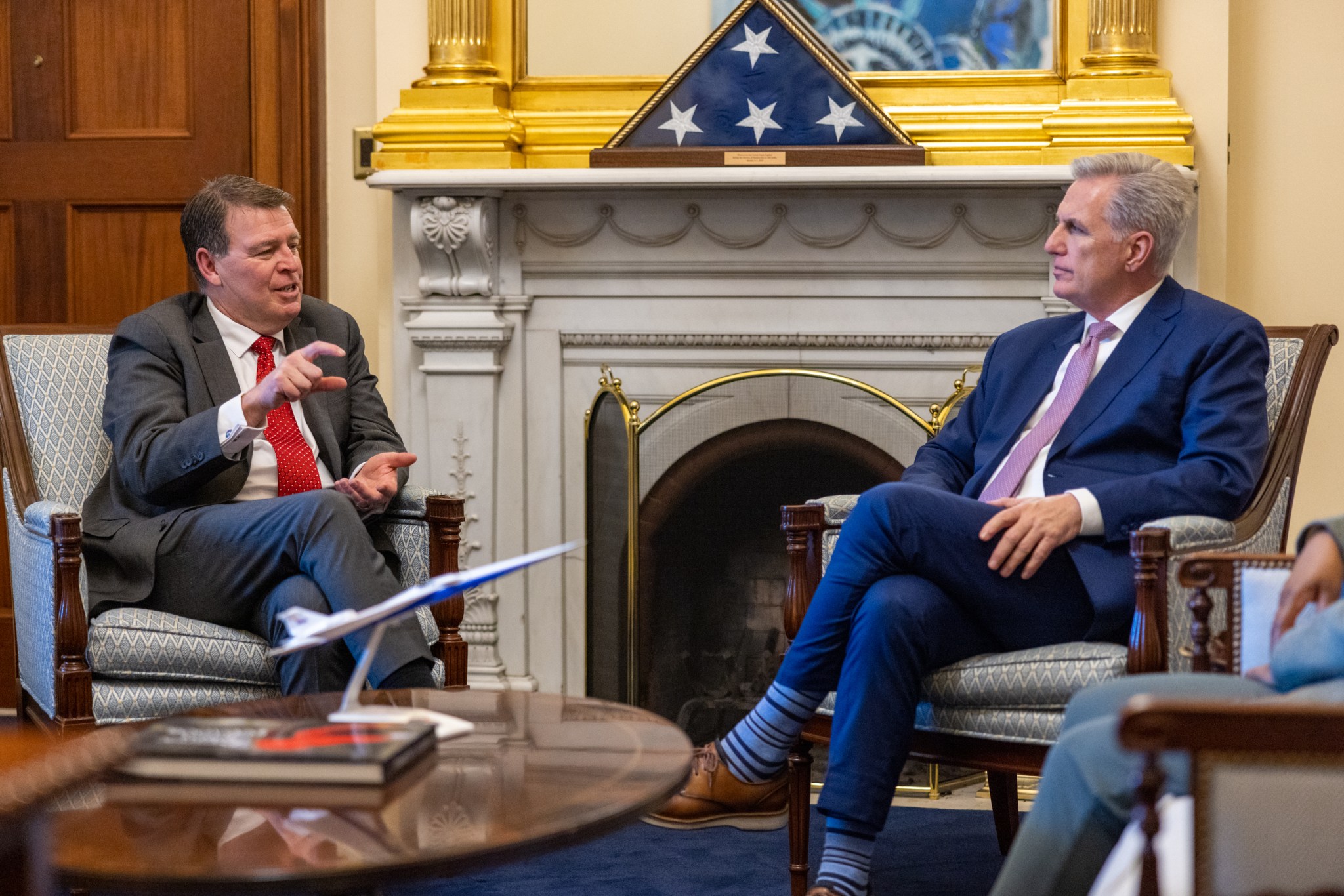 Two gentlemen are seated in front of a stone fireplace. On the mantle is an encased American flag and a golden frame for artwork not in view. In front of them is a wooden oval coffee table, where a model of NASA's X-59 is placed. The gentleman on the left, Center Director Brad Flick, is talking and motioning with his hands, wearing a great suit and red tie. The gentleman on the right, Speaker Kevin McCarthy, is listening facing Mr. Flick, wearing a blue suit and pale colored tie.