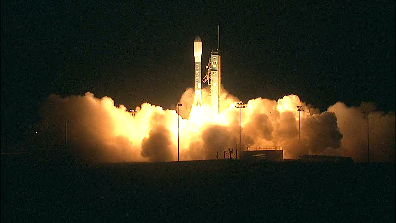 A rocket launching at night. The rocket is illuminated by its own flame, which also lights the steam clouds below it gold.