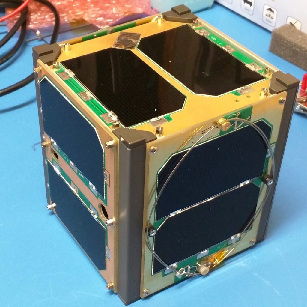 ELaNa 14 JPSS-1 CubeSat RadFxSat sitting on a blue table. The cubesat is gold and black, with gold making up the framework and smooth black on each side of the cube.